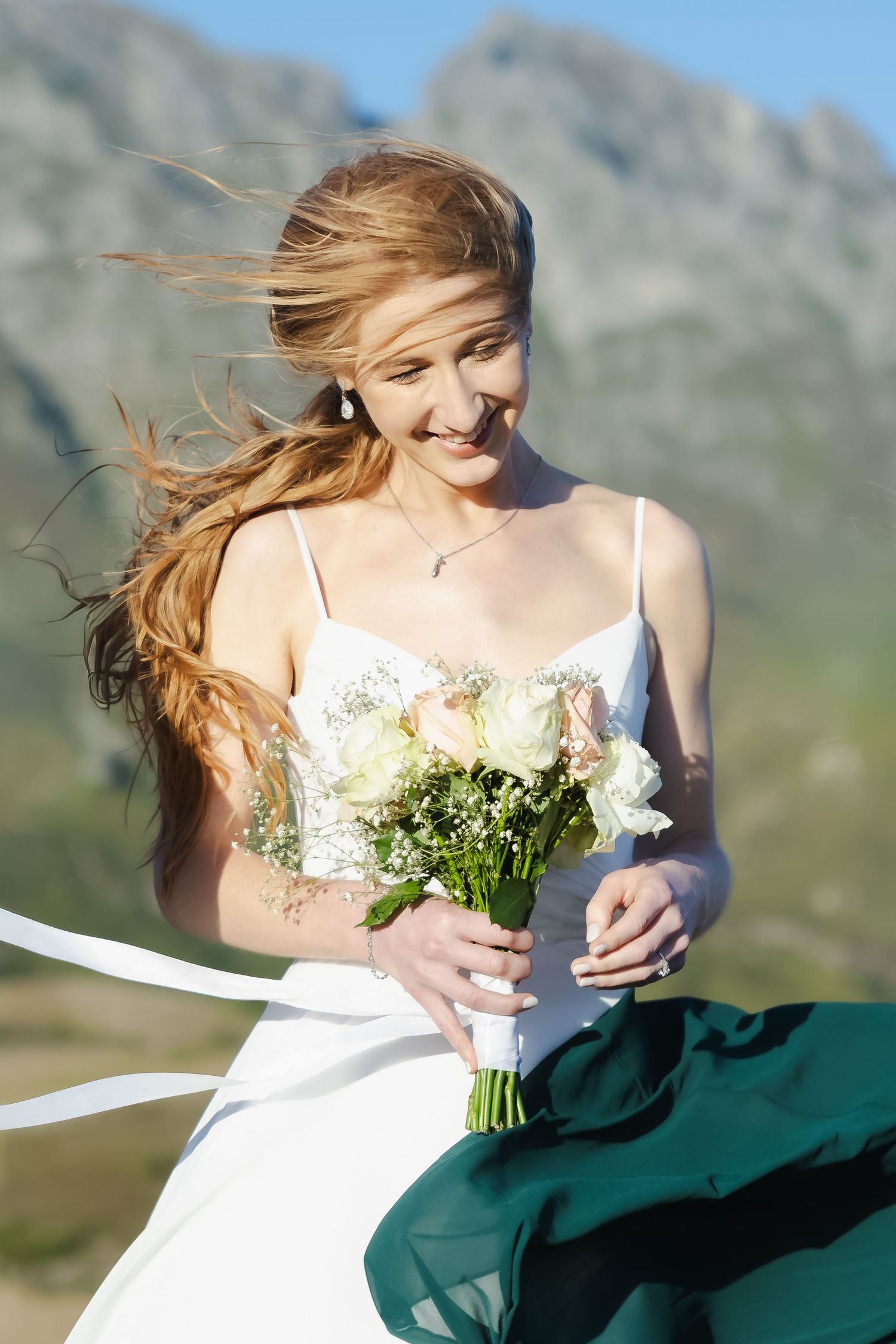 A person in a white dress holds a bouquet against a mountainous backdrop as wind blows their hair.