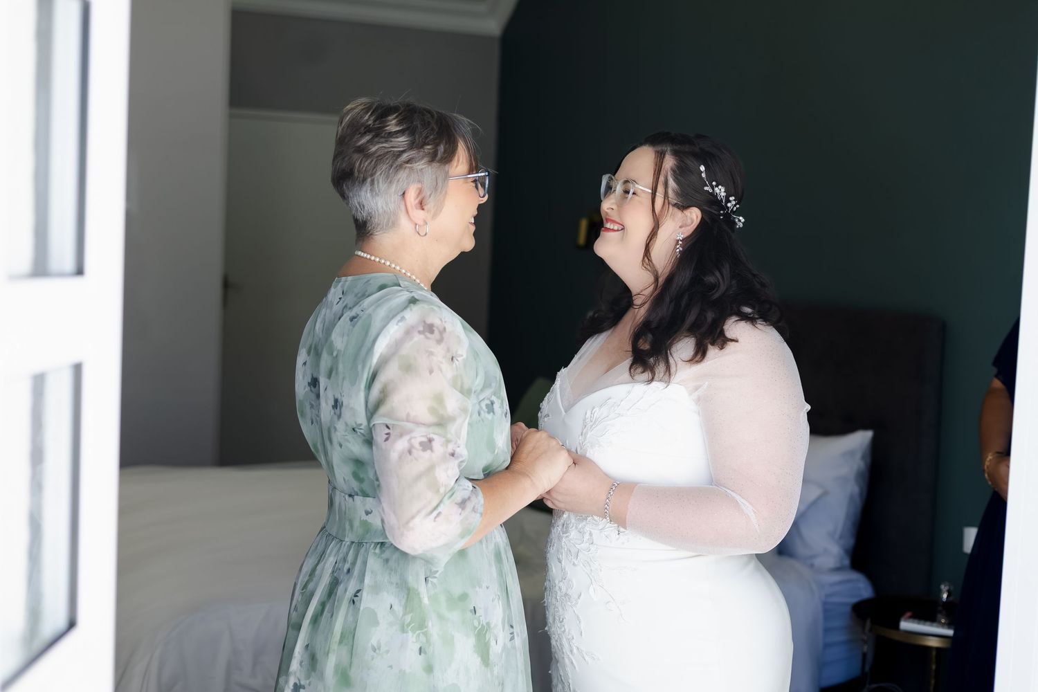 Bride in white lace gown shares a tender moment with older woman in floral dress in a green room.