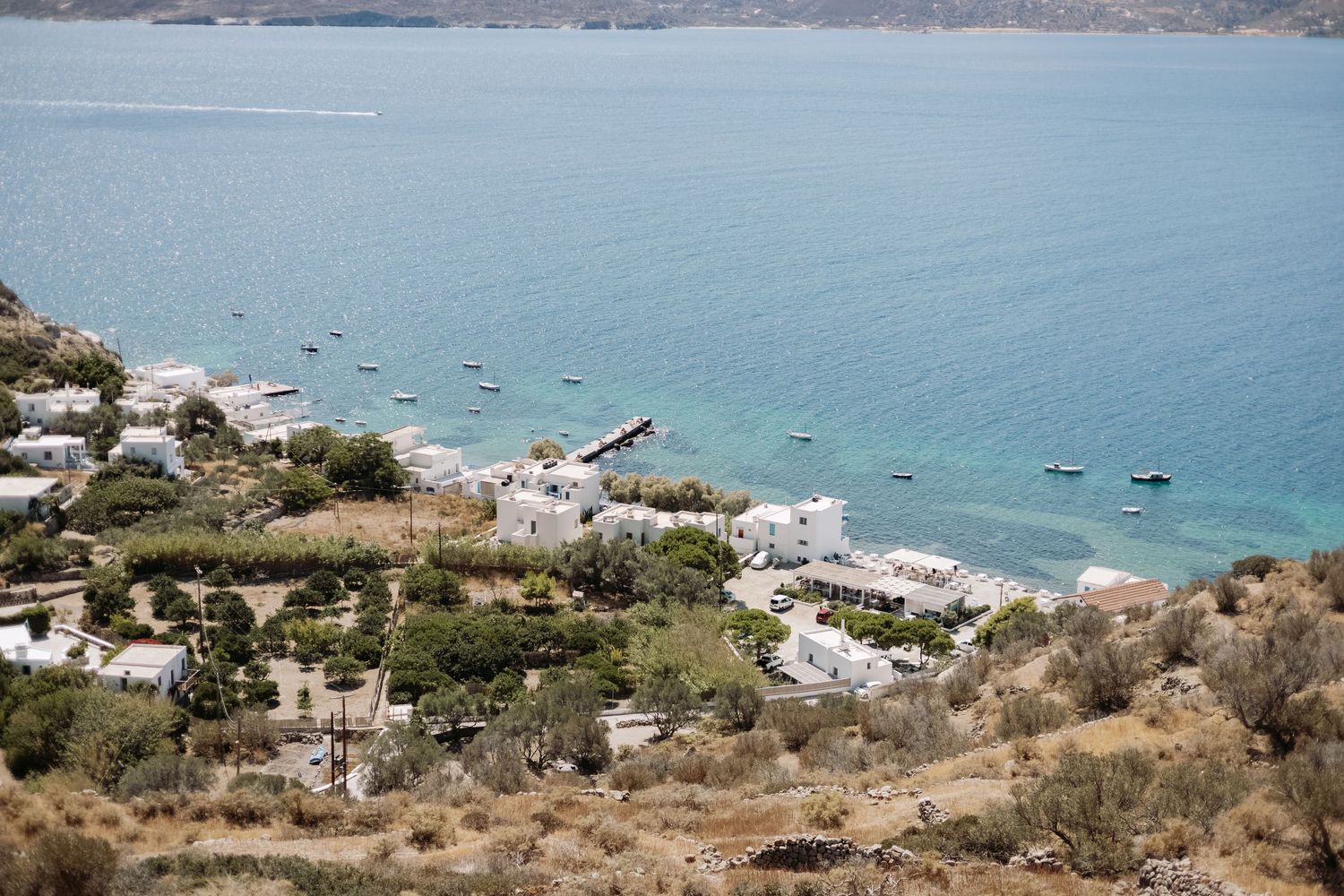 Aerial view of a small Greek coastal village with white buildings, boats in turquoise water, and rocky hillside terrain.