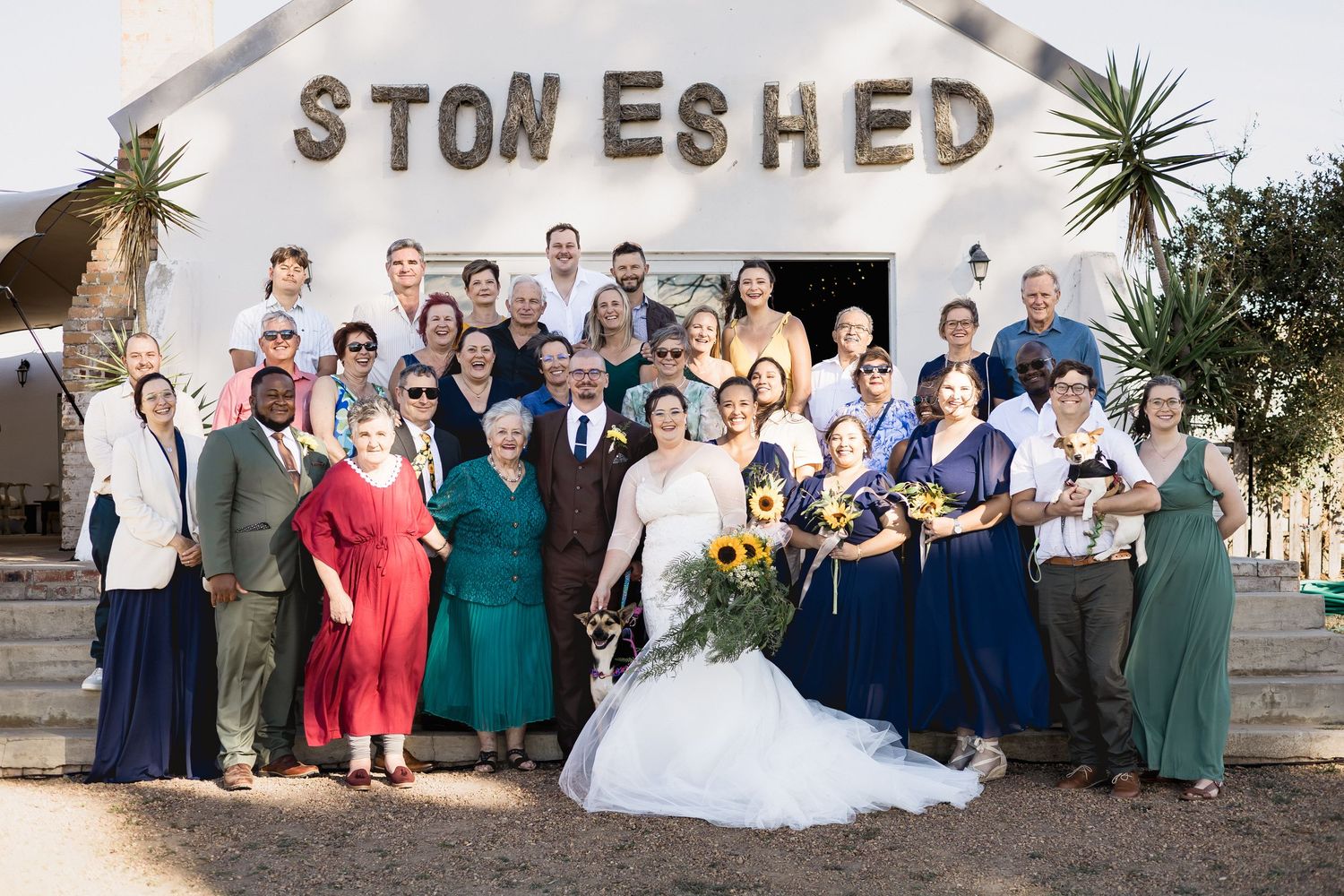 Wedding party posing in front of Stone Shed venue, bride in white gown with sunflowers, groom and guests in formal attire.