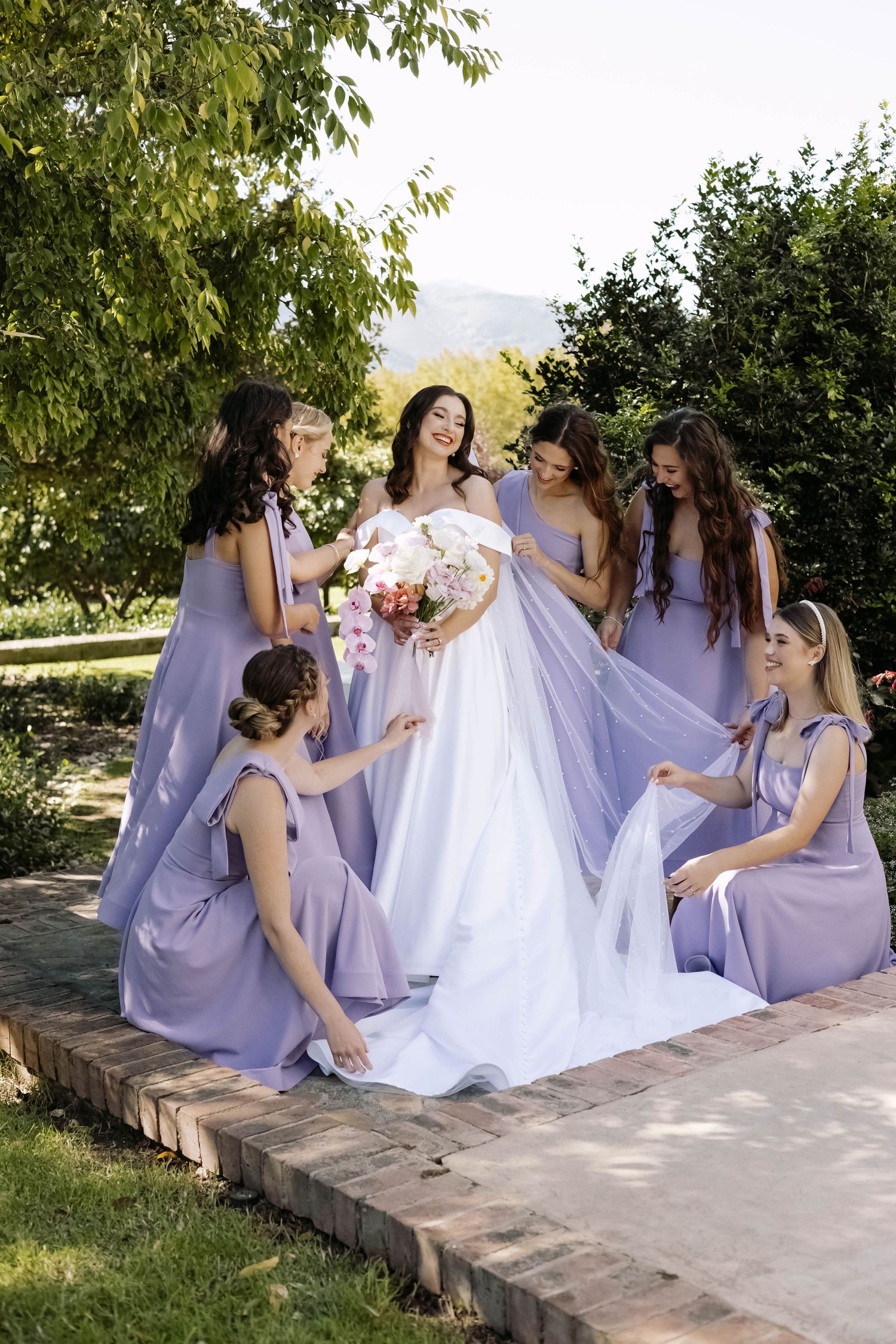 A group in light purple bridesmaid dresses gather around a white wedding dress on stone steps in an outdoor setting.
