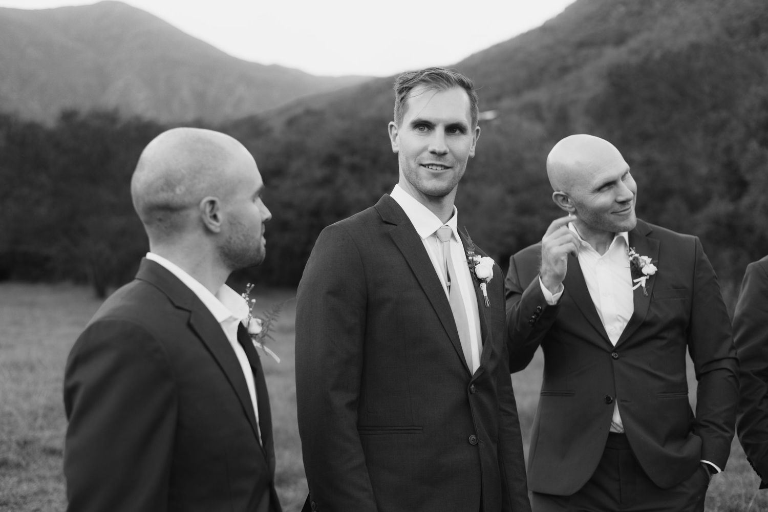 Groomsmen in black suits share laughs against a scenic mountain backdrop.