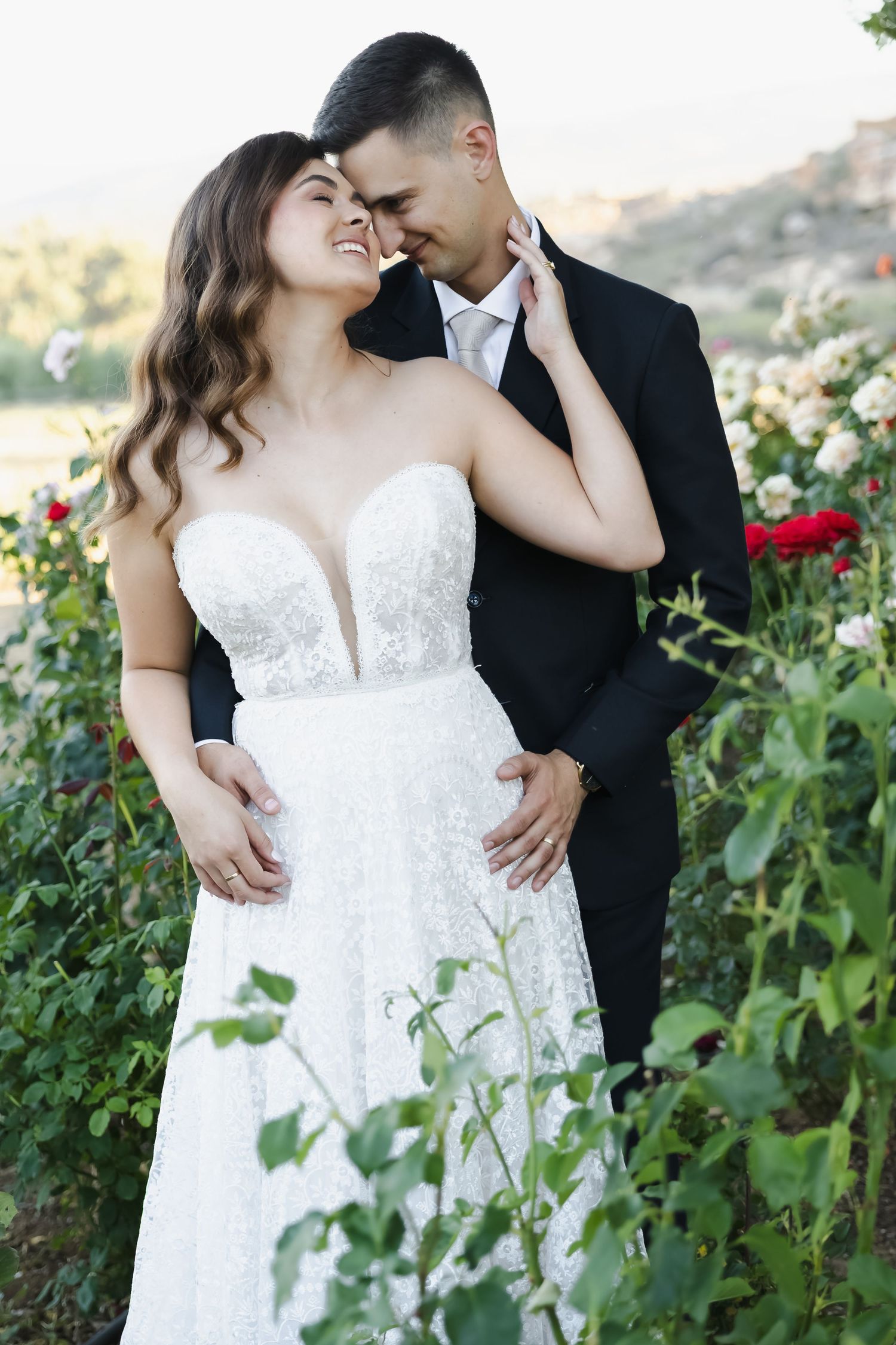 Romantic wedding couple embracing among red roses in a garden during golden hour sunset.