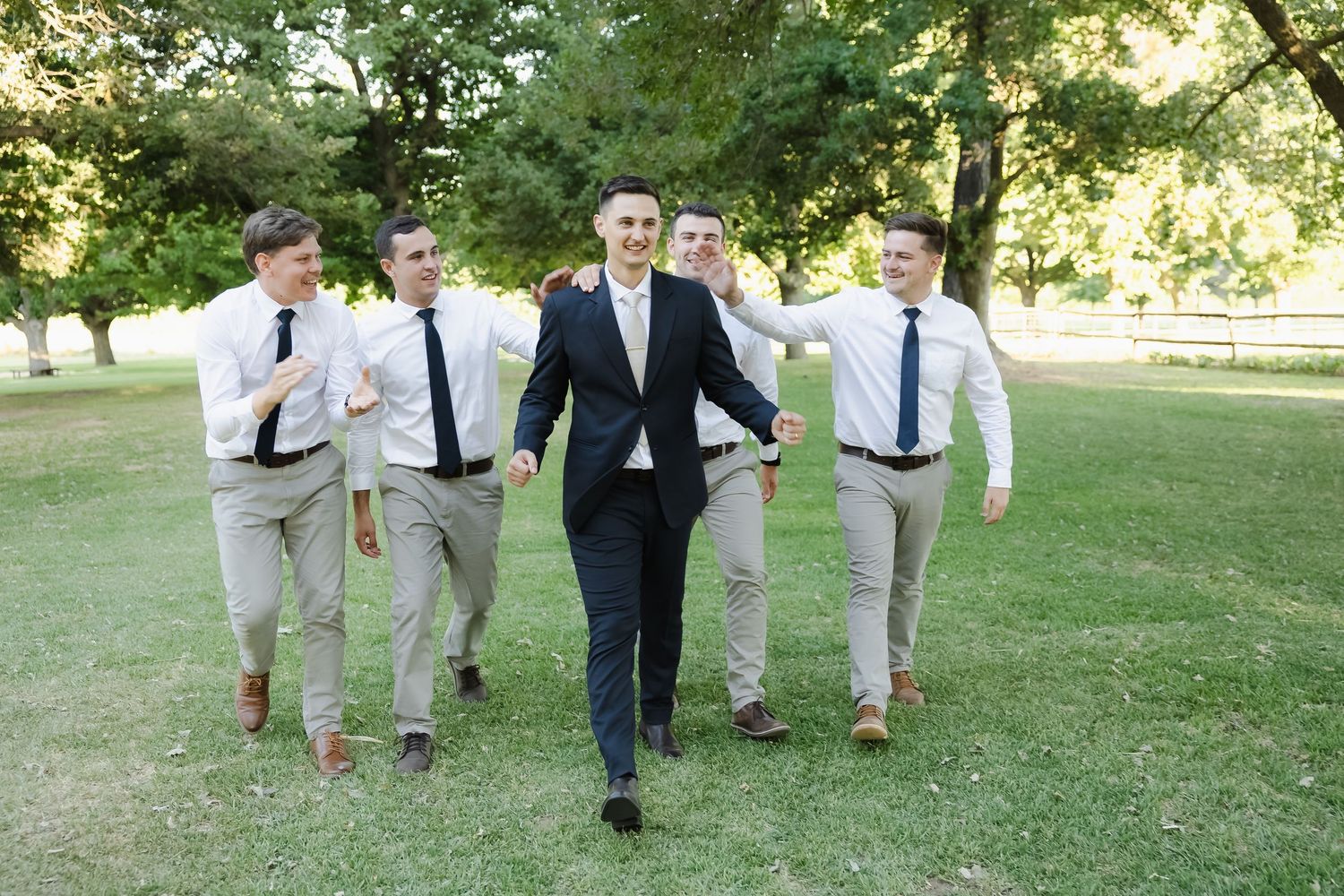 Groomsmen in light gray suits and blue ties walk together on a lush green lawn during a summer wedding celebration.