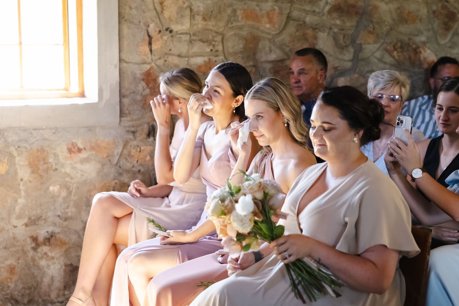 Bridesmaids in pink dresses sitting together and reacting emotionally during a wedding ceremony.