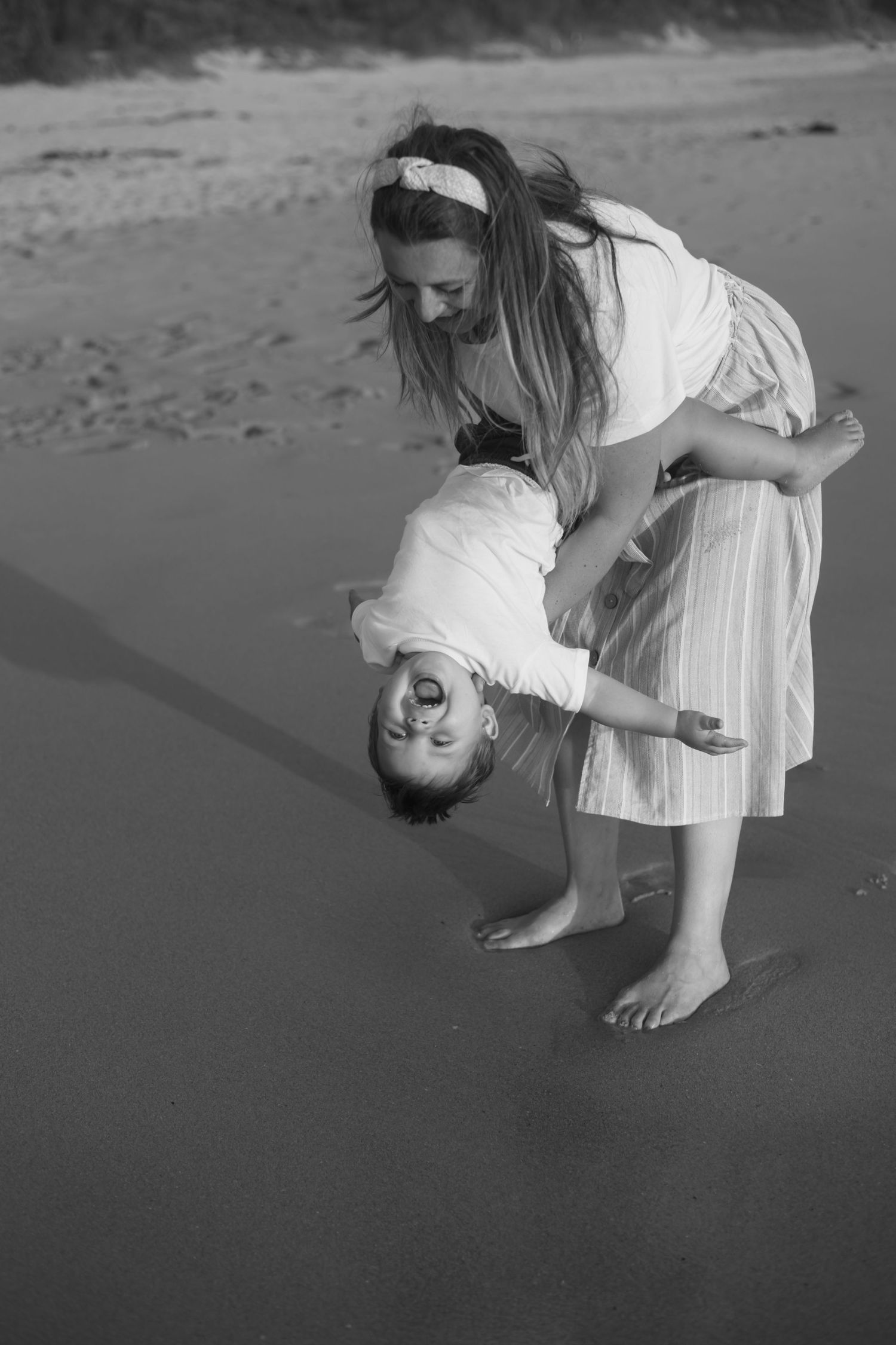 Black and white photo of a mother playfully holding her child upside down on a sandy beach.