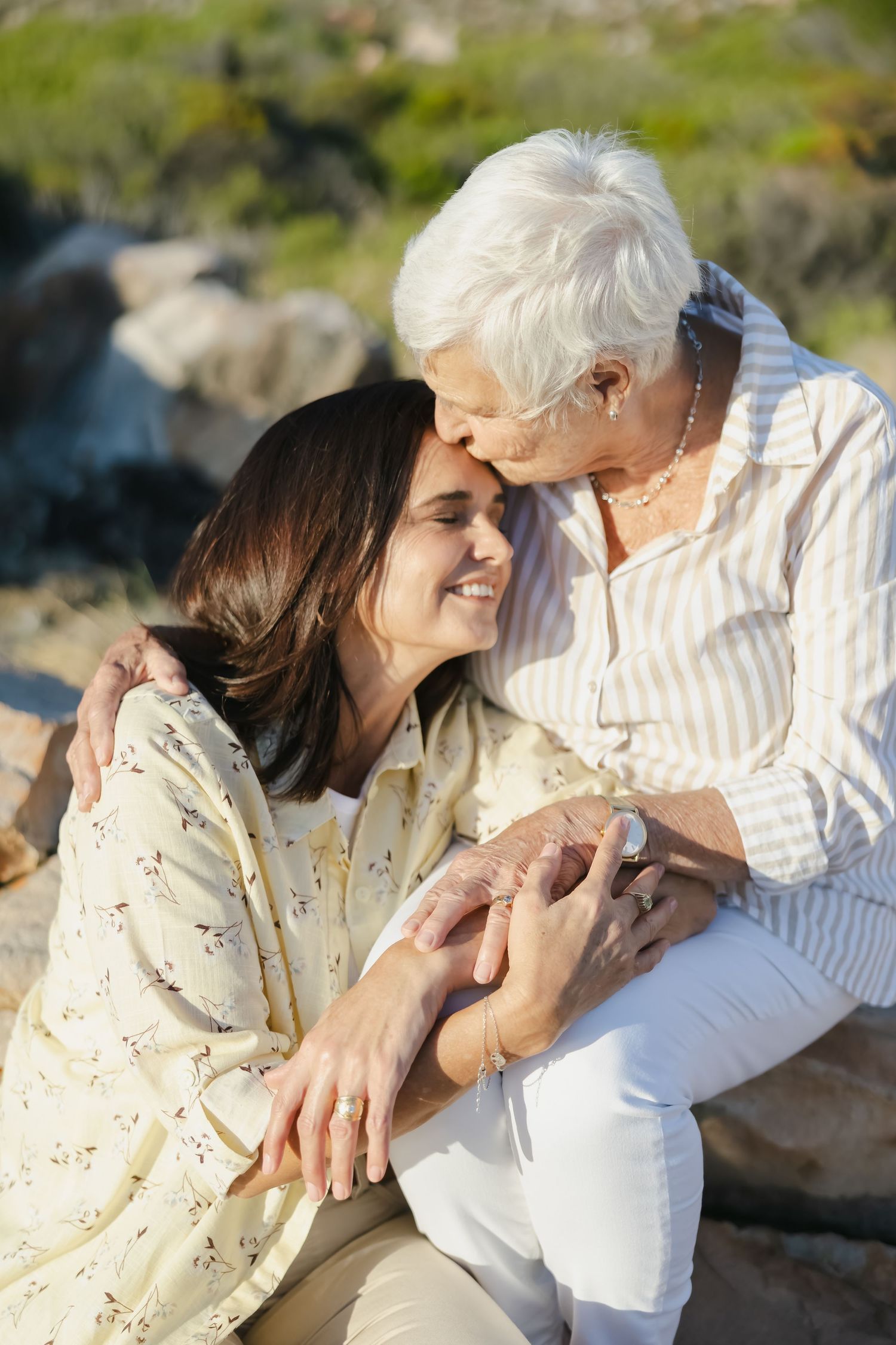 Senior woman kissing younger woman on forehead outdoors, sharing a tender mother-daughter moment near rocky landscape.