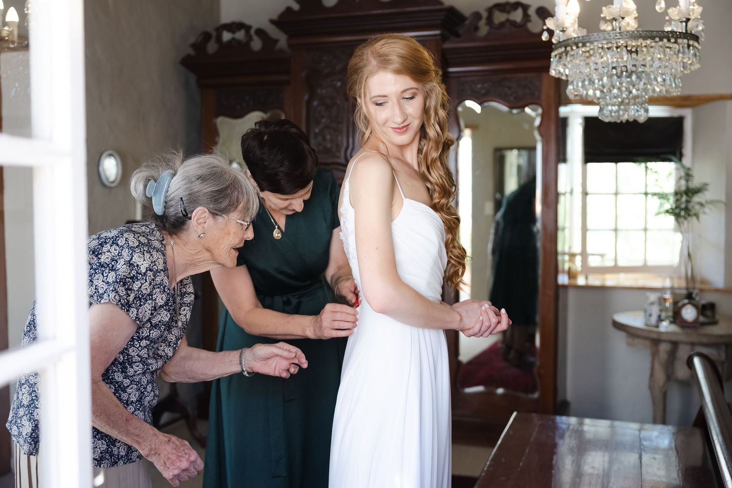 A bride gets ready in an elegant room with a crystal chandelier as helpers assist with final preparations.