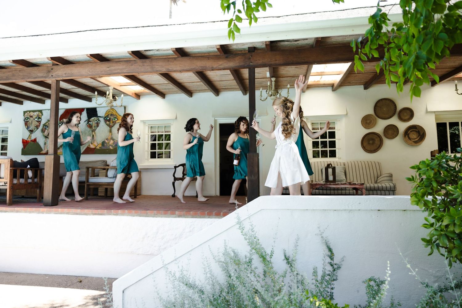 Wedding party gathers under an elegant outdoor pergola with hanging decorations and greenery.