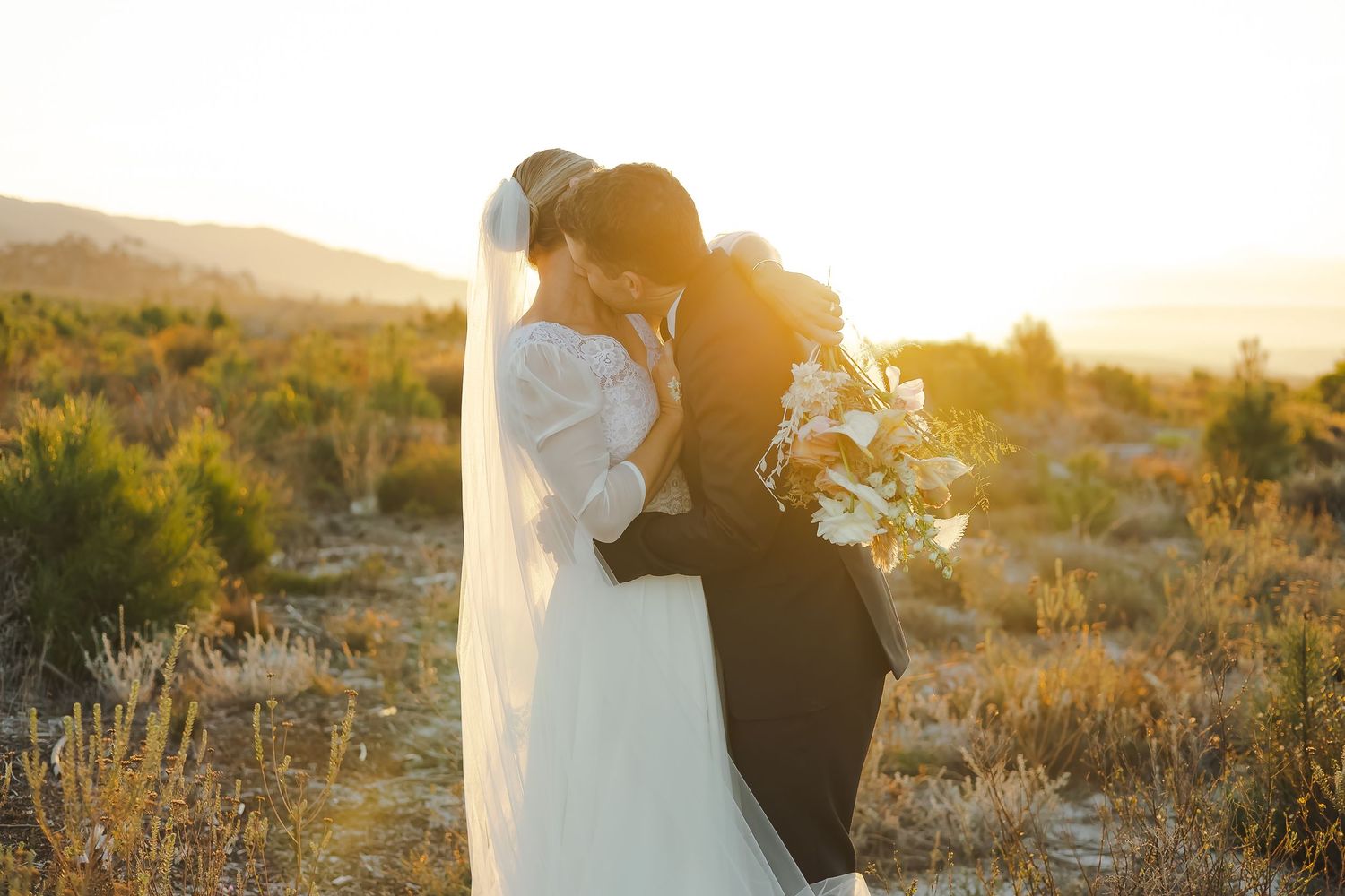 Wedding couple photographed in golden hour light among desert brush and mountains.