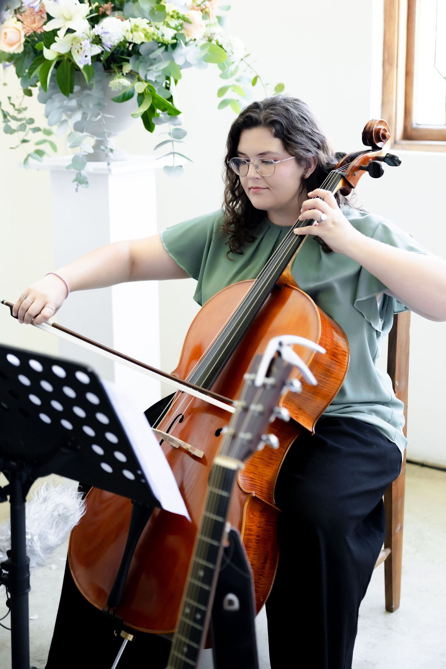A musician plays the cello next to a music stand in a bright room decorated with floral arrangements.