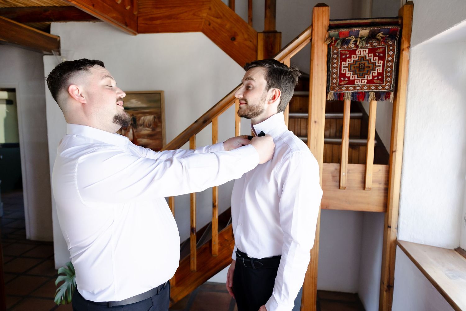 Two people in white shirts engage in playful interaction near a wooden staircase in a home interior.