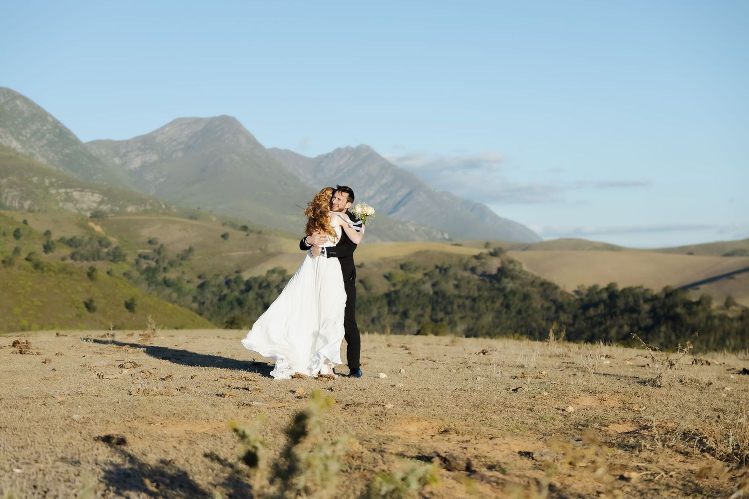 A couple in wedding attire embraces while standing on a scenic mountain hillside with rolling peaks in the background.