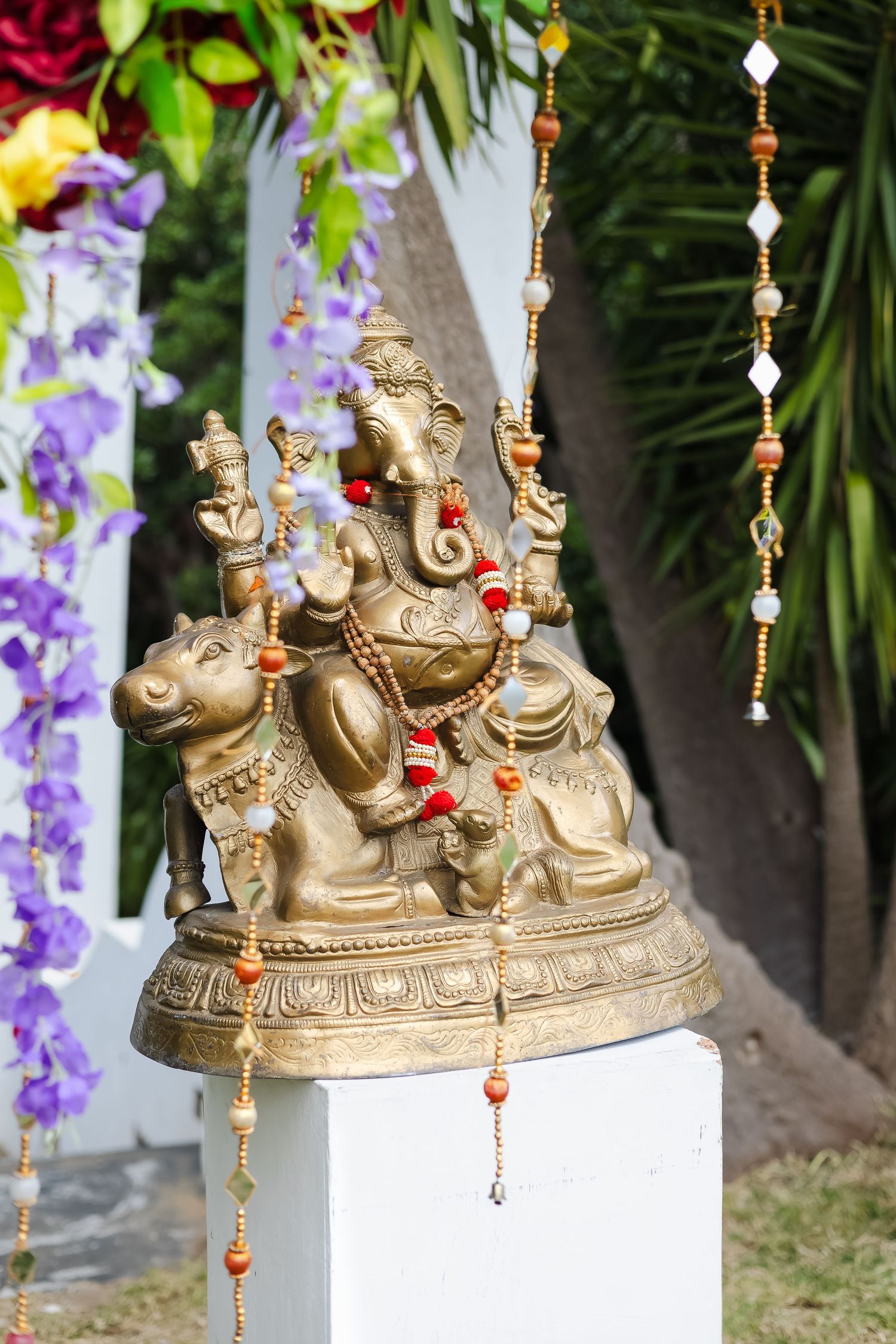 Decorative brass Ganesh statue with beaded hanging ornaments displayed against purple flowers in a garden setting.