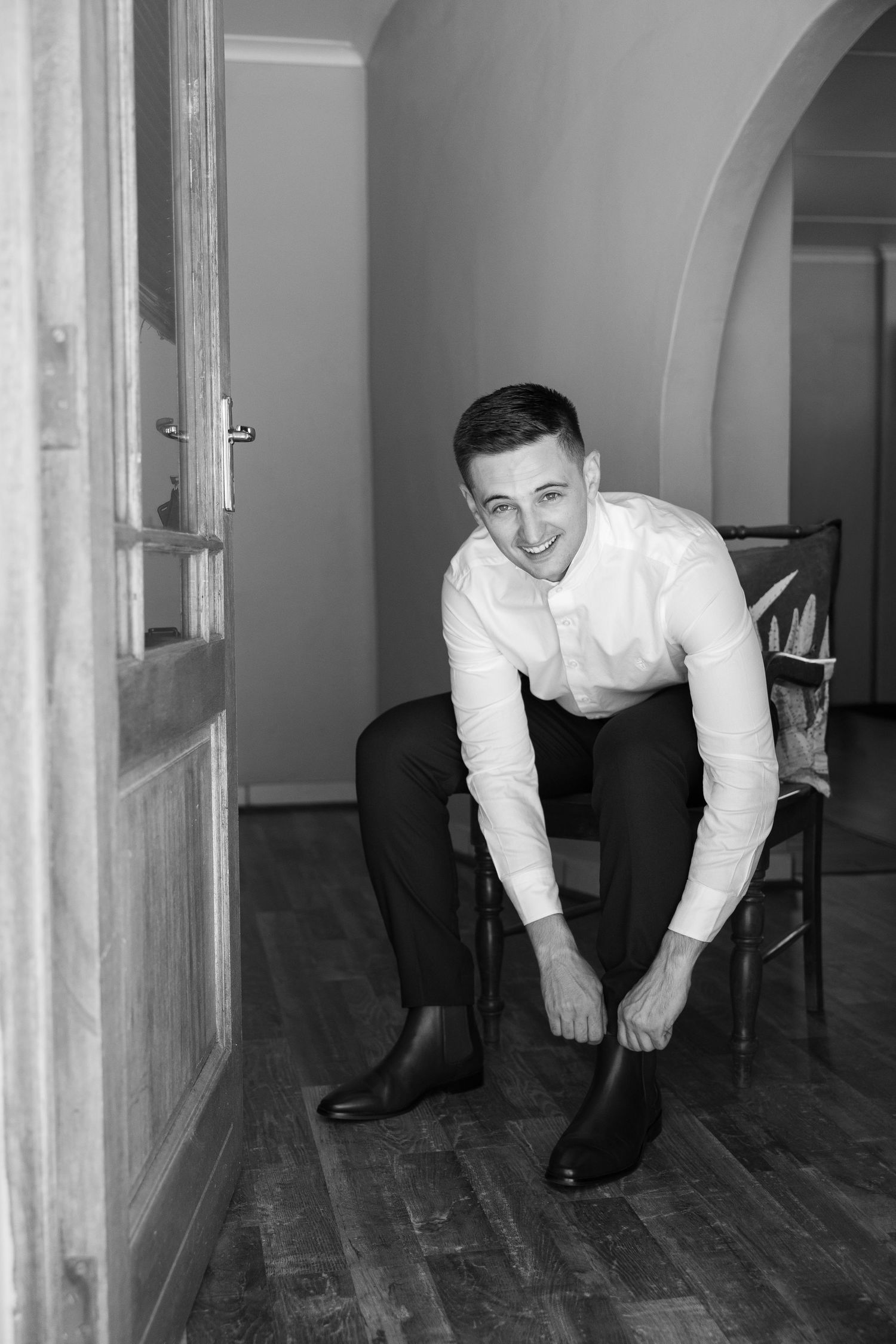 A person in a white dress shirt and dark pants sits on a chair while putting on socks in a room with wooden floors.