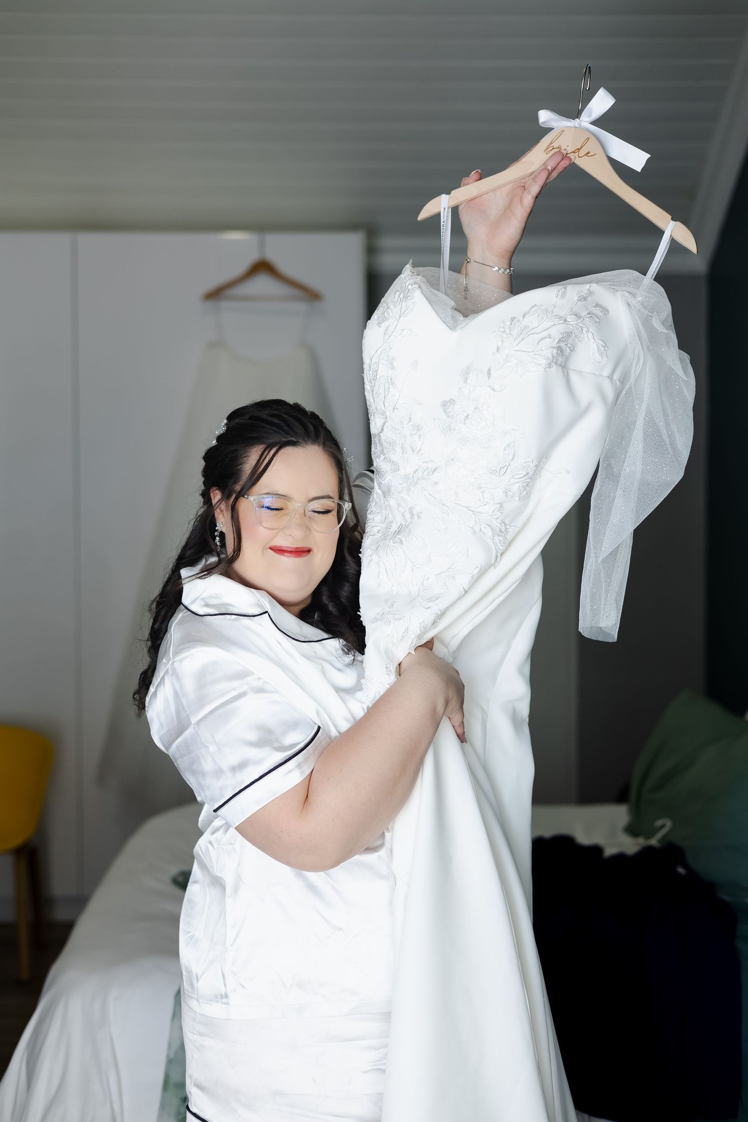 Bride with Down syndrome in white pajamas holds up her wedding dress on a hanger in a bright bedroom.