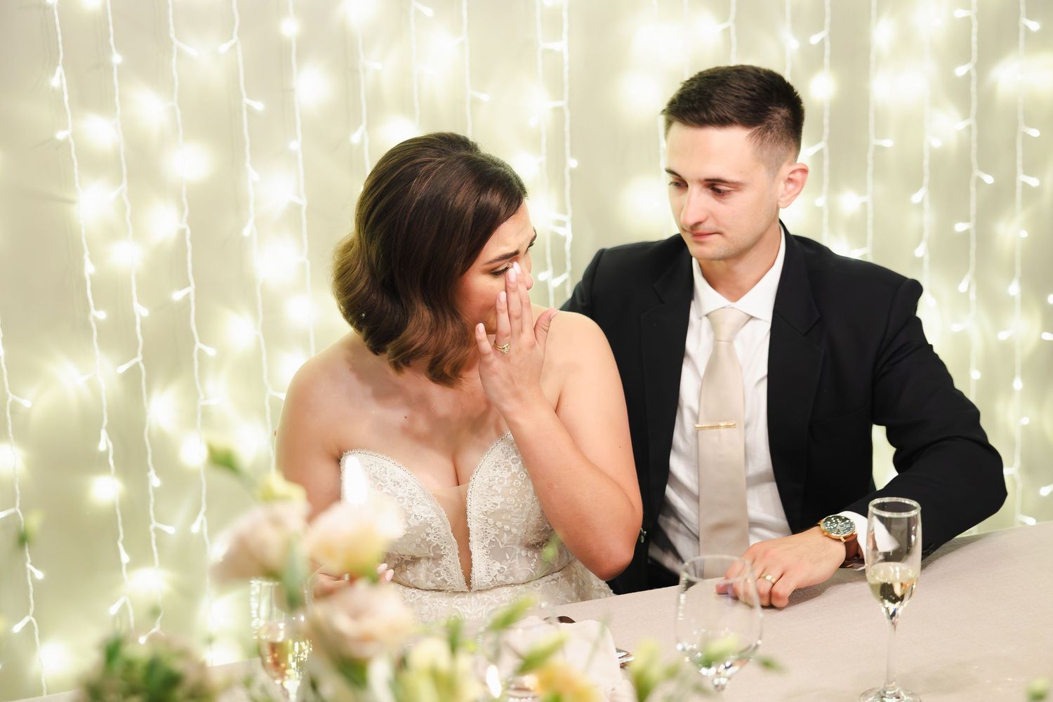 Wedding reception scene with twinkling fairy lights in background as couple sits at decorated table.