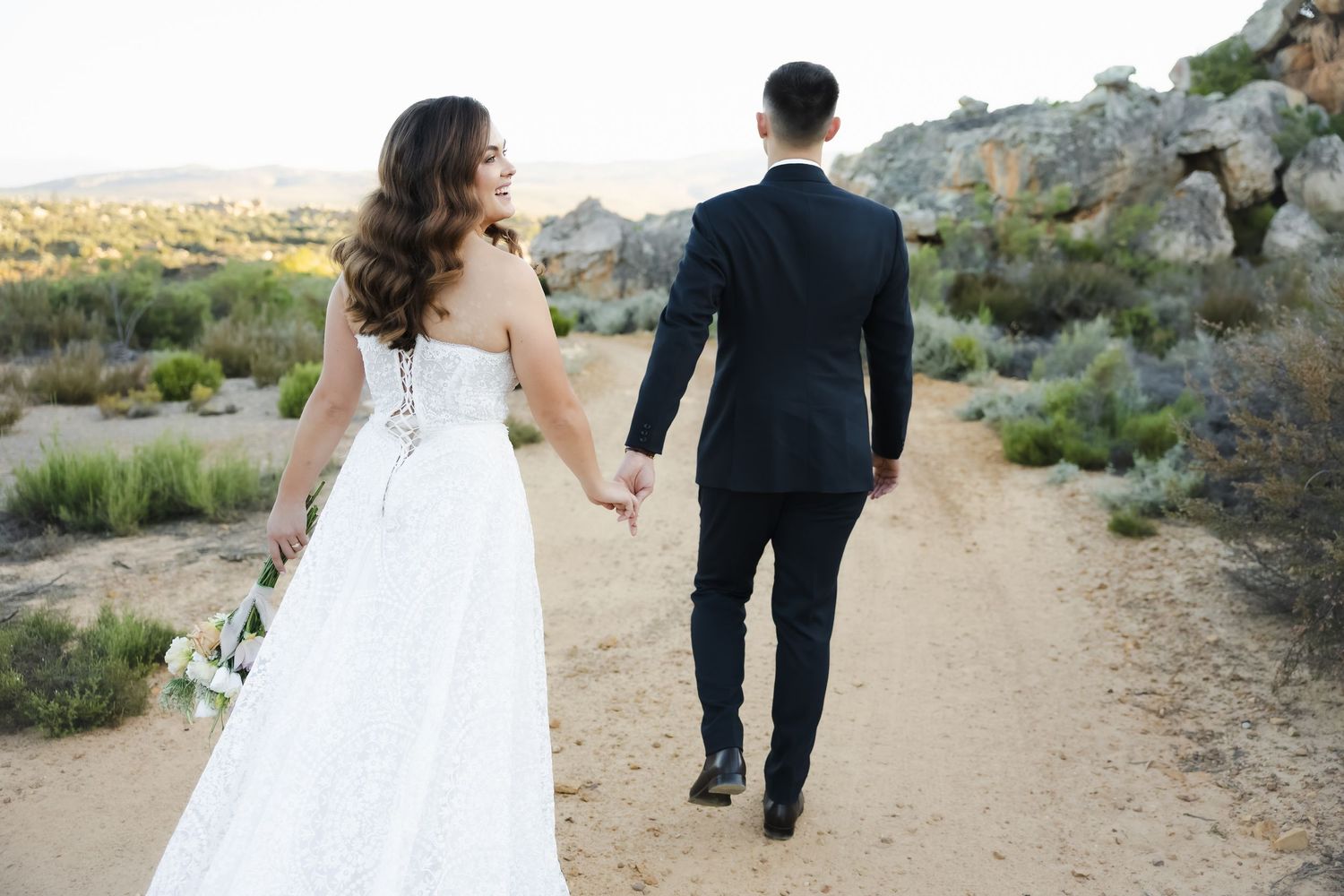 A couple walks hand in hand on a desert trail, with the bride in a white gown and groom in a black suit.