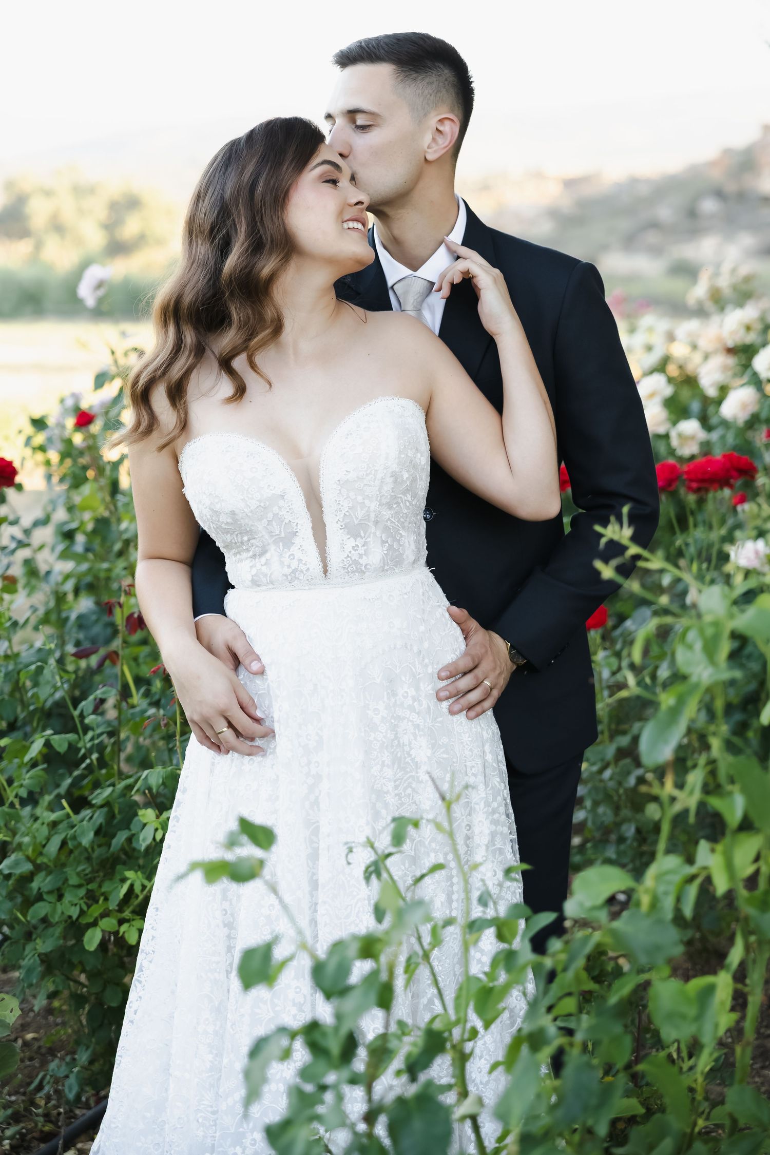 Romantic couple embraces in garden setting with white dress and black suit amid blooming roses at sunset.