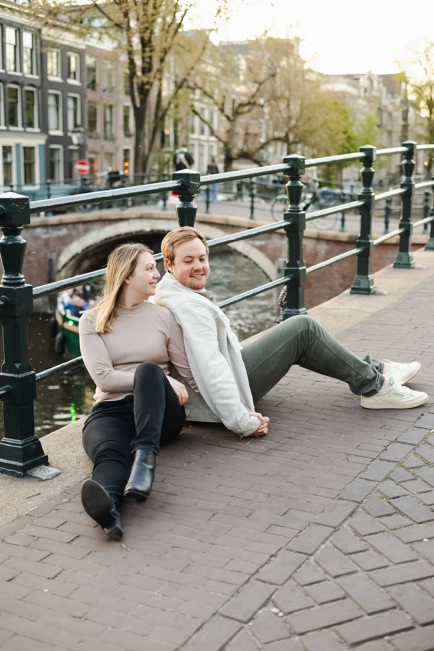 Couple sitting together on Amsterdam canal bridge, smiling, with historic buildings and waterway in background.