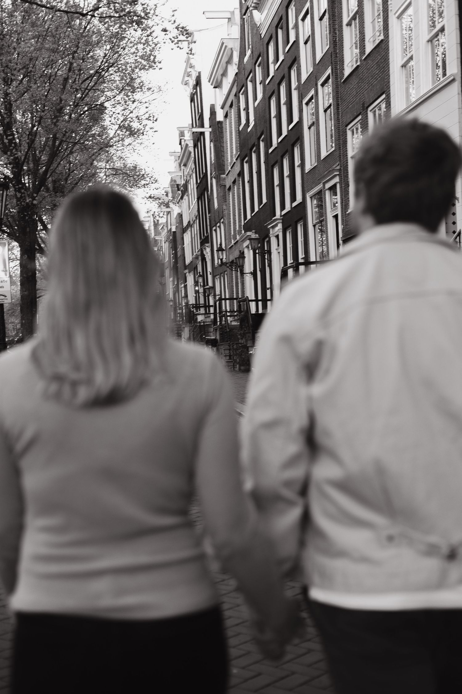 Black and white photo of a couple holding hands while walking along a historic Amsterdam canal street.