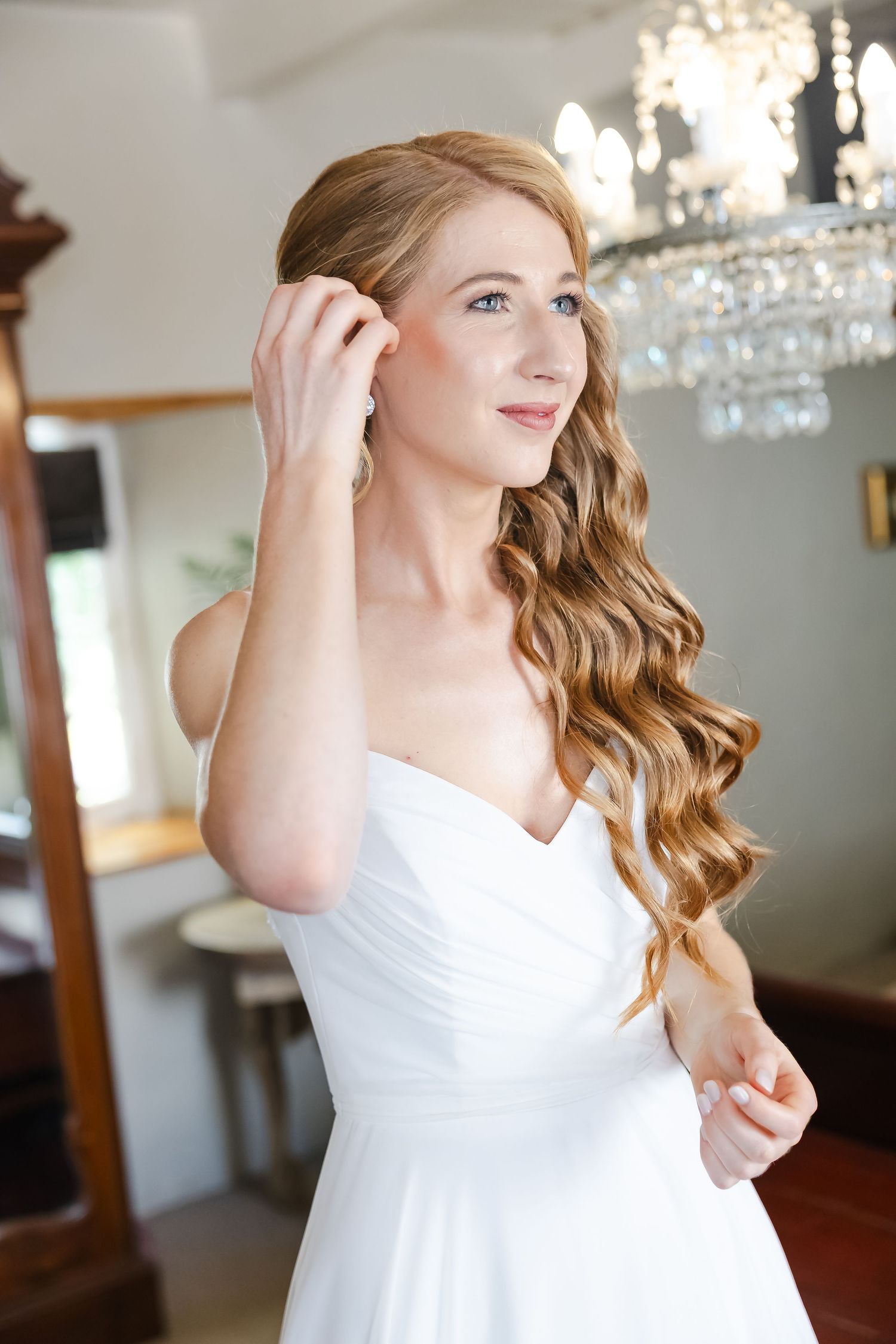 A bride in a white wedding dress with flowing curled hair poses in front of a crystal chandelier.