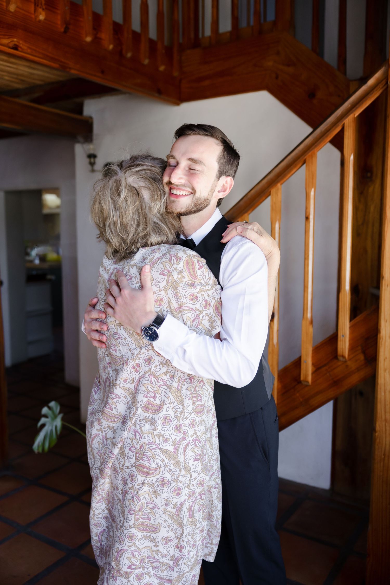 Person in formal vest shares warm embrace with someone in floral dress by wooden staircase.
