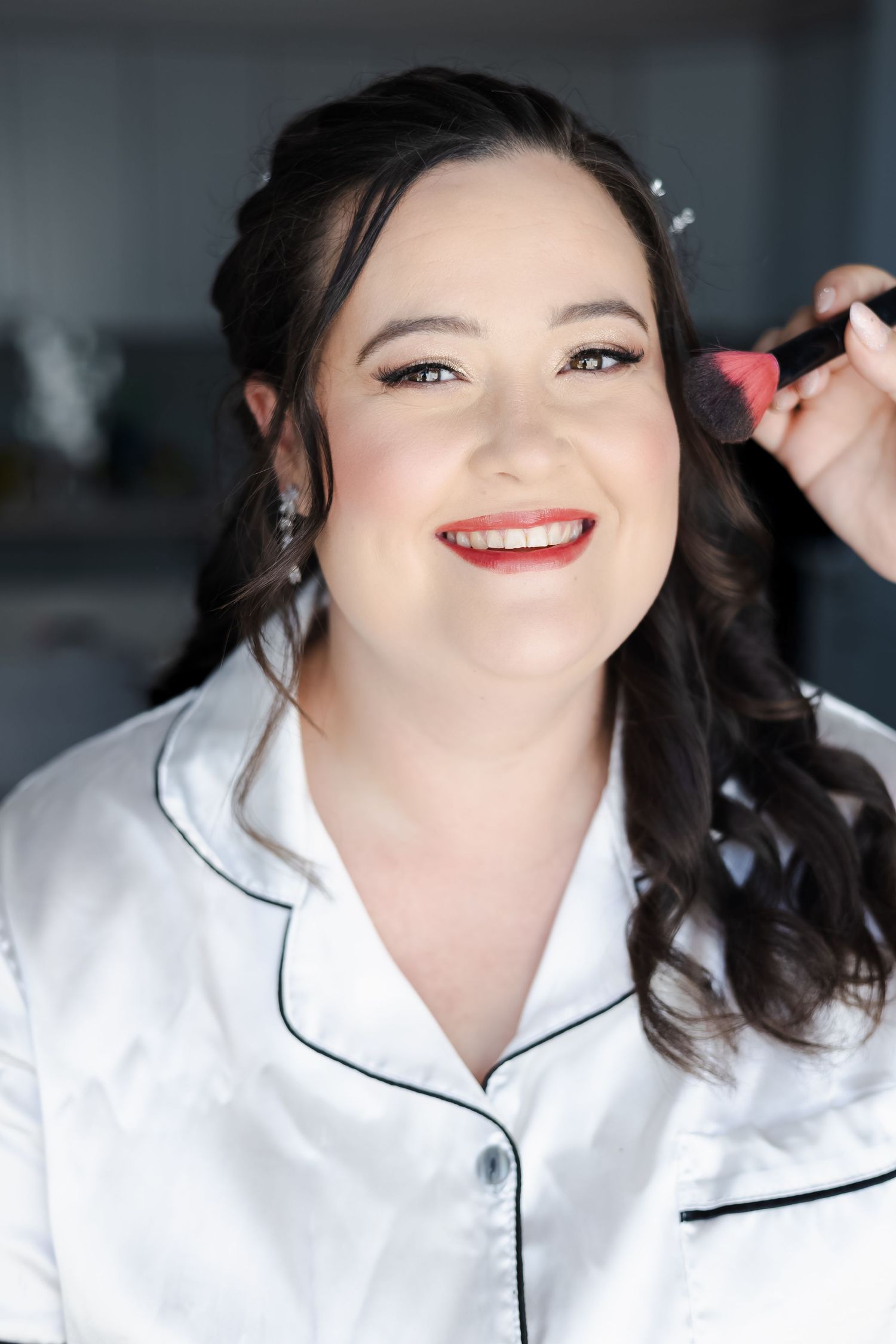 Smiling bride getting blush applied with a makeup brush, wearing white satin pajamas and red lipstick.