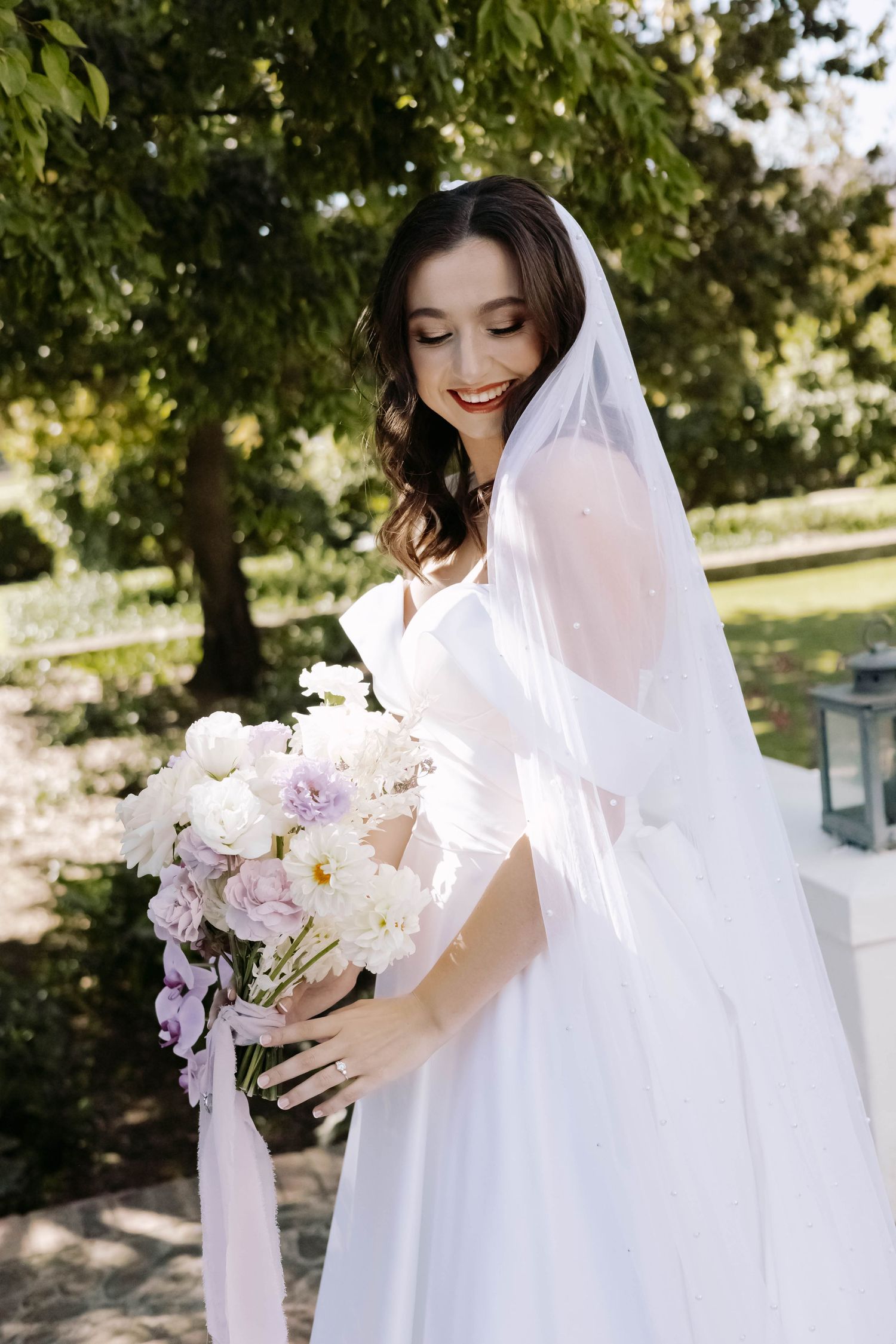 A bride in a white wedding gown holds a purple and white orchid bouquet in a garden setting.