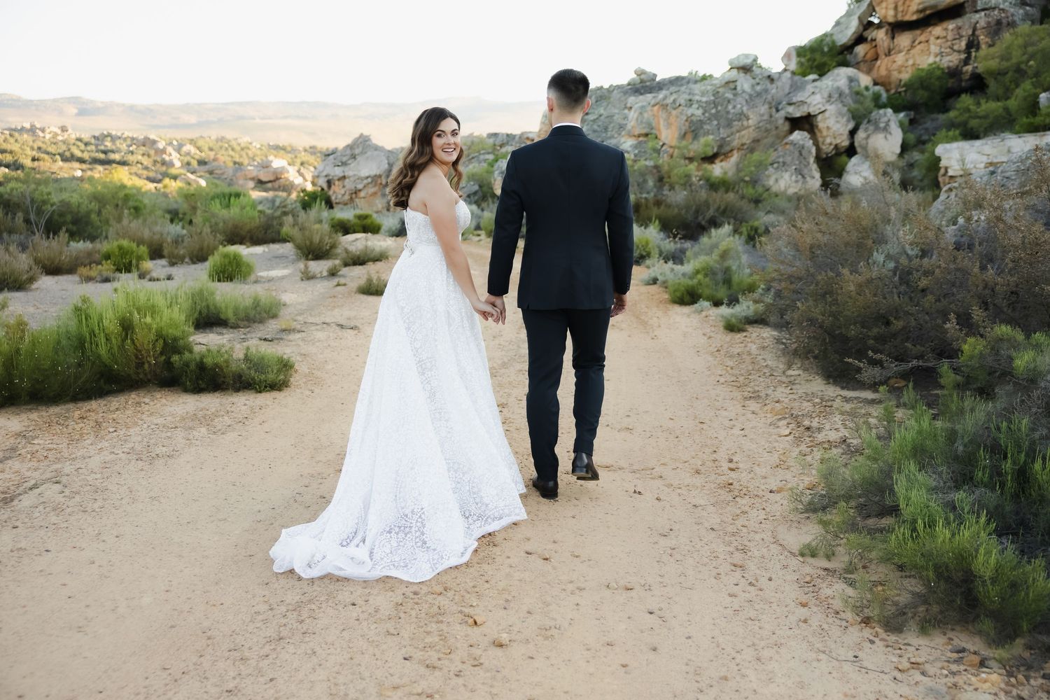 A couple in wedding attire walks along a desert trail surrounded by desert brush and scenic mountain views.