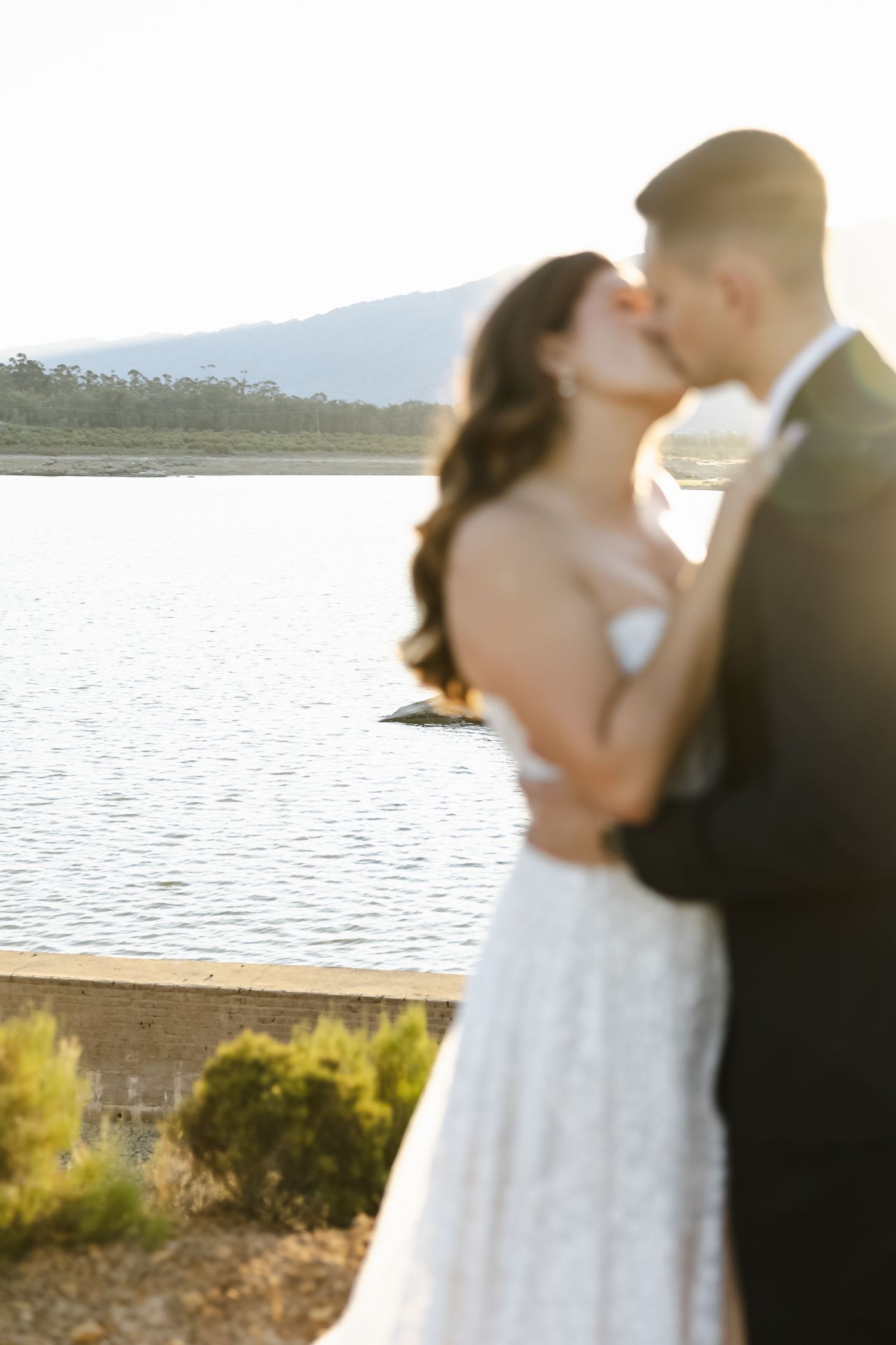 A romantic sunset kiss between newlyweds by a serene waterfront.