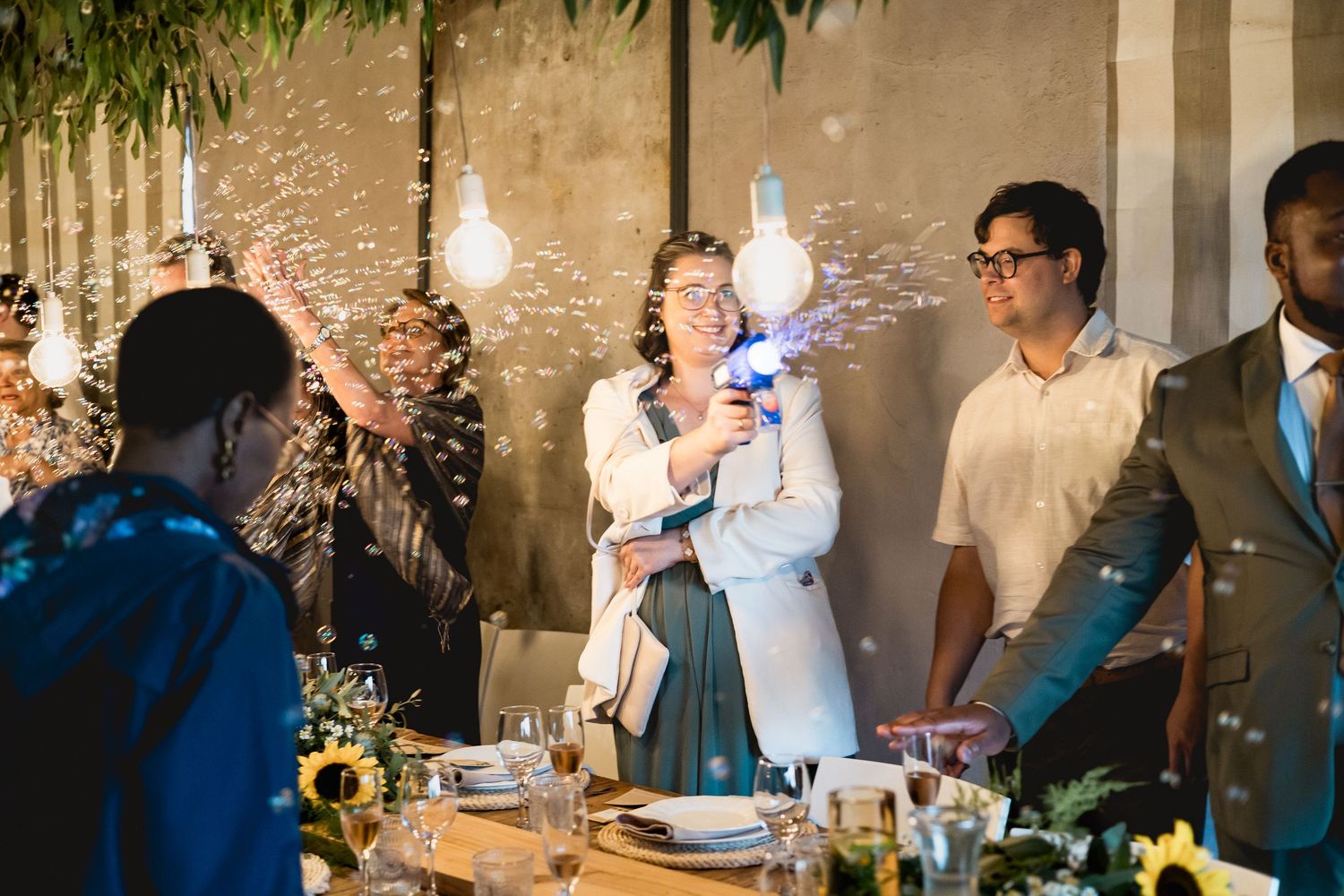 Woman shoots bubble gun at festive indoor party with sunflowers and Edison bulb decor while guests celebrate.