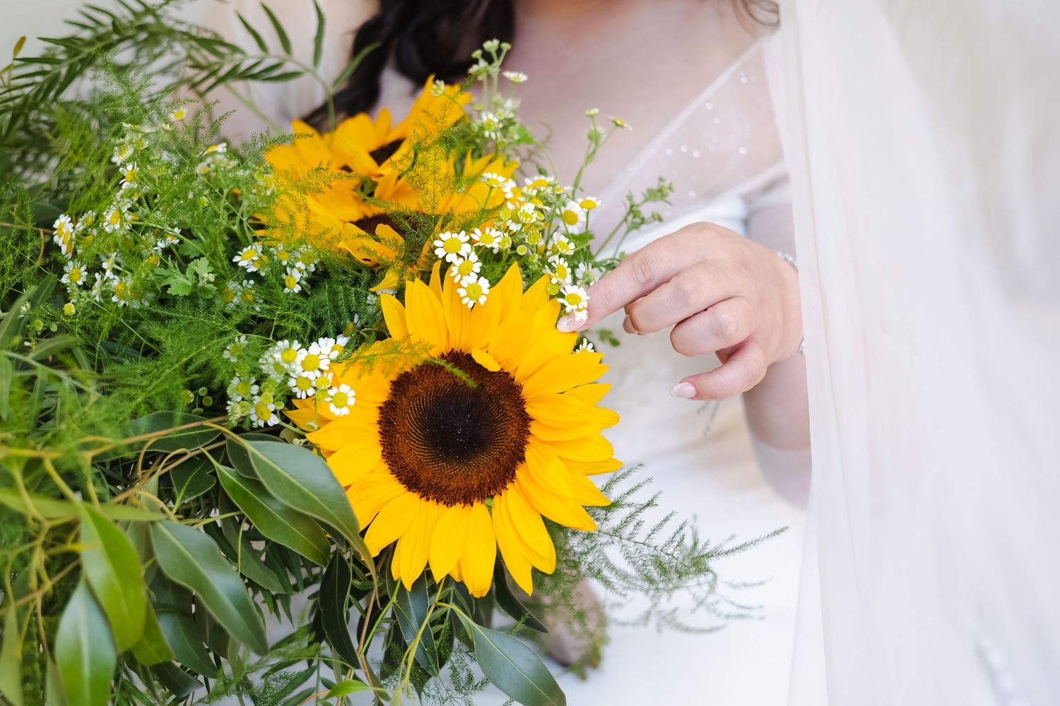 Bride holding a vibrant sunflower wedding bouquet with wildflowers and greenery, wearing a white lace gown and veil.