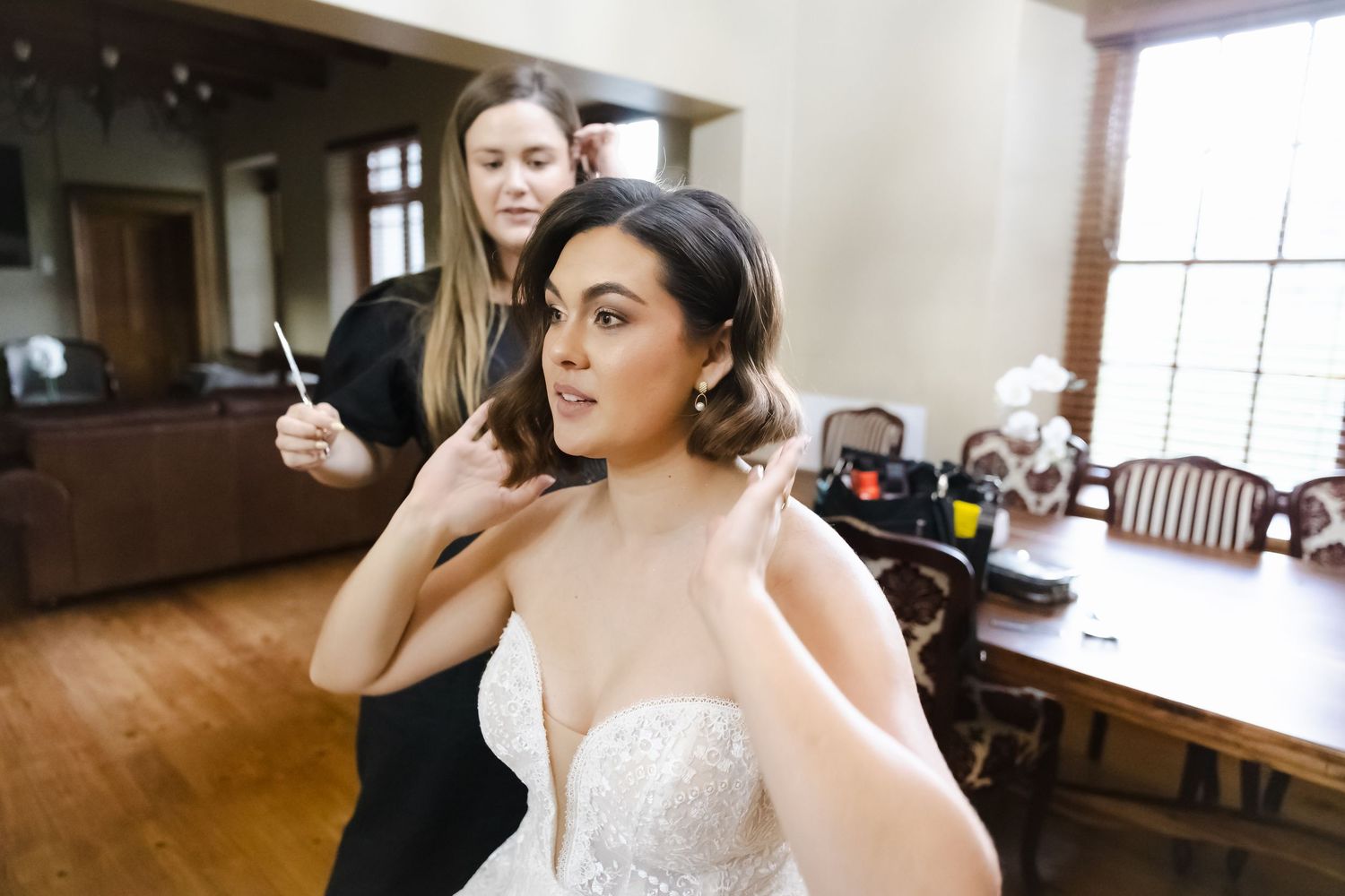 A bride in a white lace wedding dress gets her hair and makeup done in a bright indoor setting.