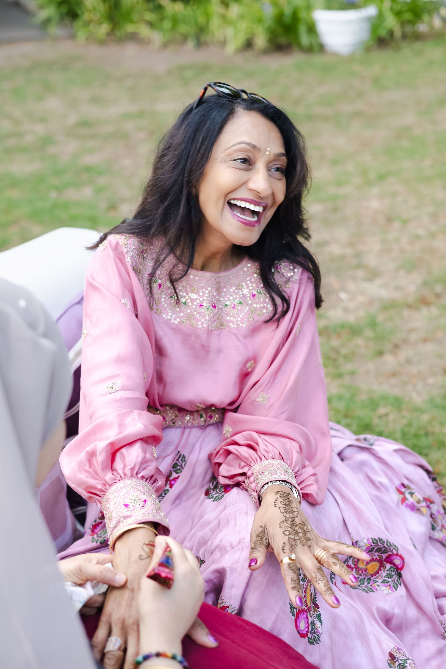 A person in a pink embroidered dress sits outdoors on grass while showing decorated hands with henna designs.