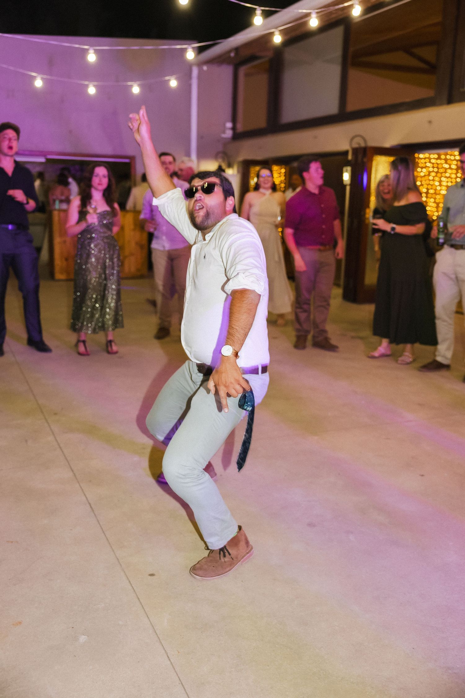 People dancing and having fun at a wedding reception with purple lighting and string lights overhead.