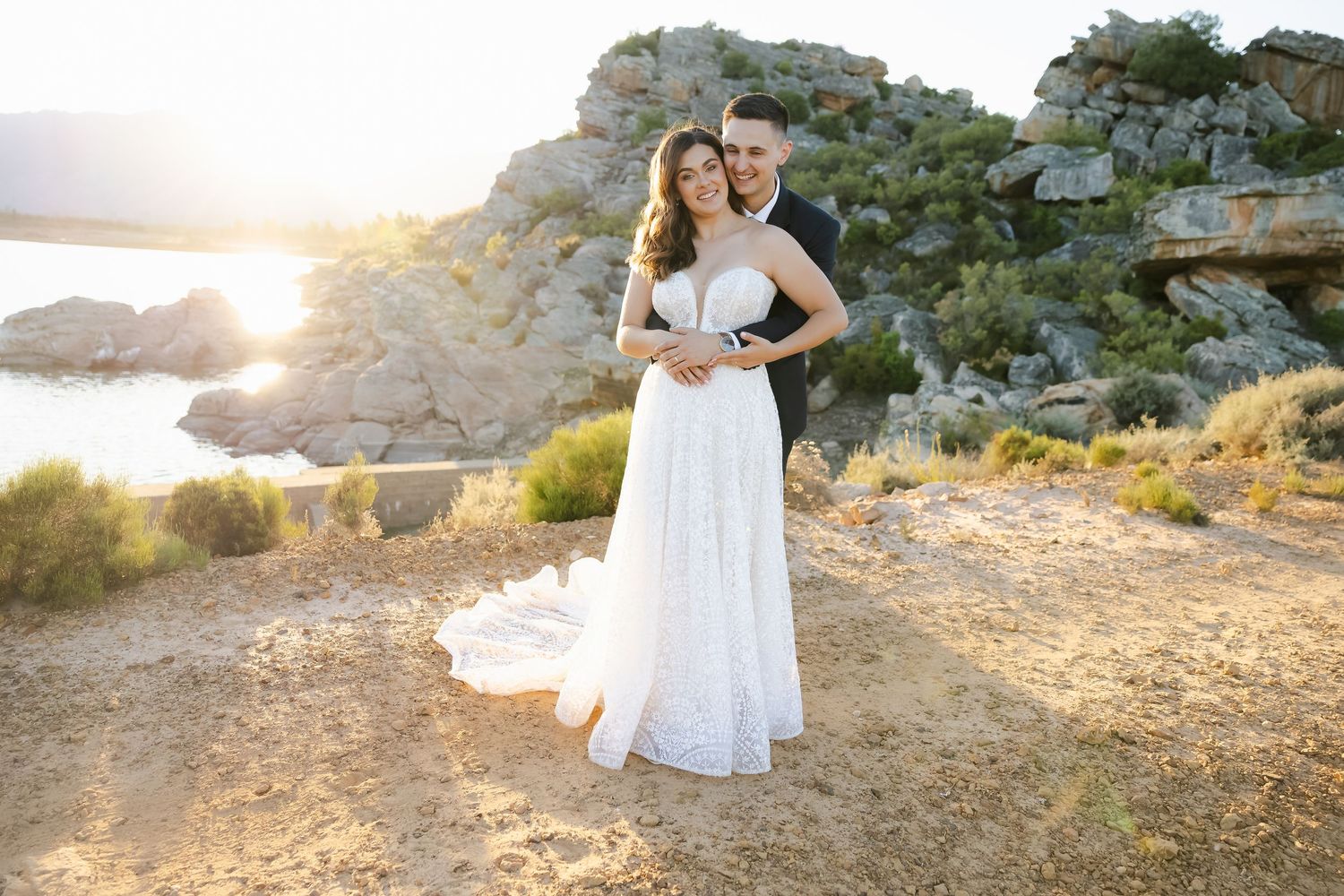 Newlyweds pose together at sunset with rocky cliffs and ocean views in the background.