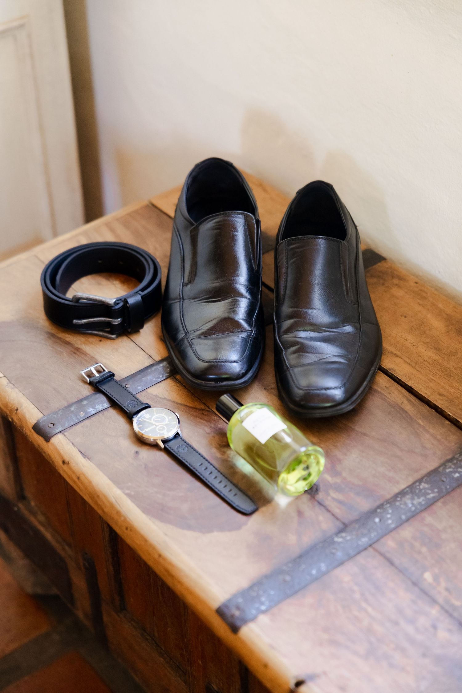 Men's formal accessories including black leather dress shoes, belt, watch and cologne bottle on wooden surface.