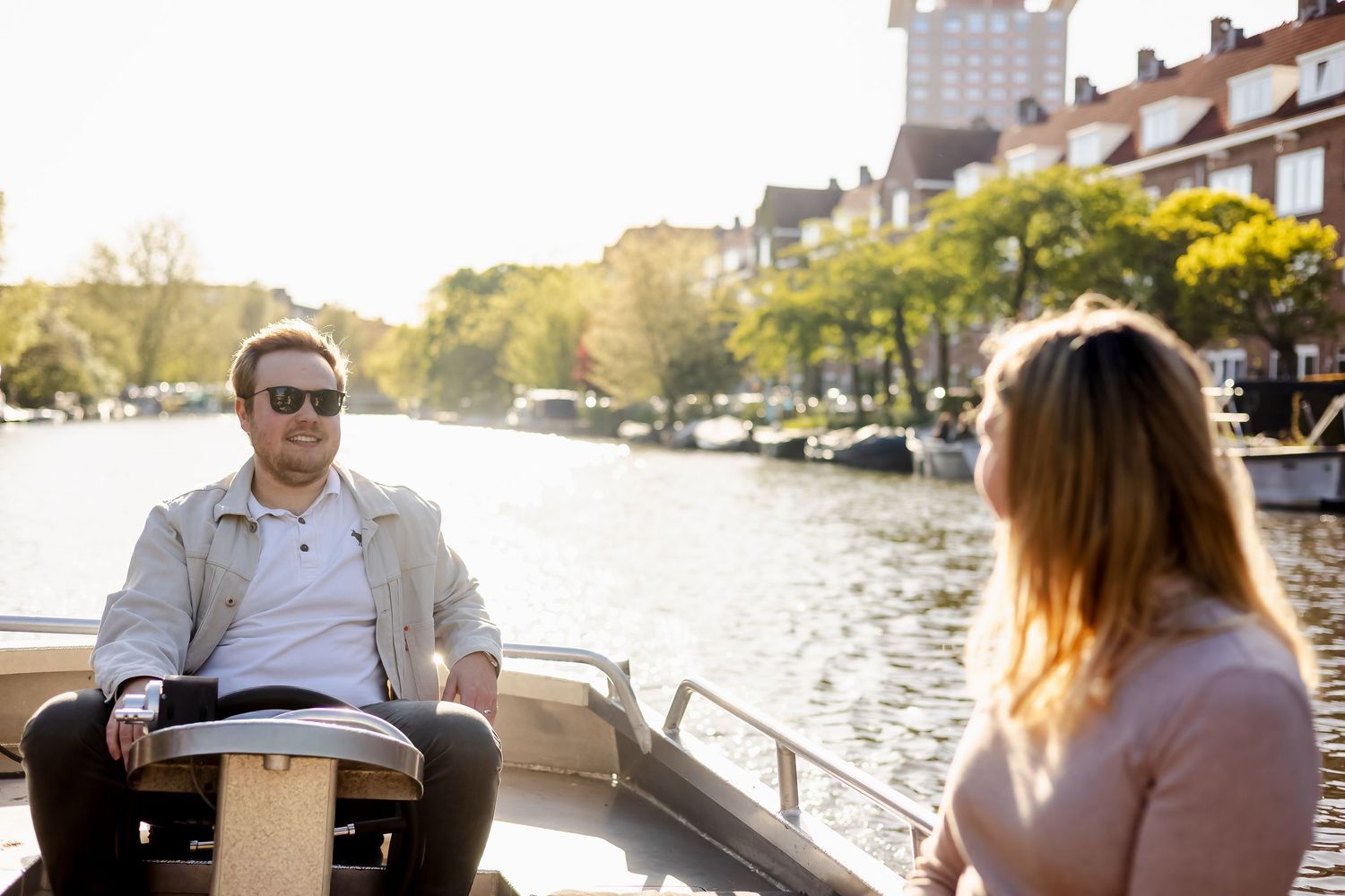 Man and woman enjoying a sunny boat ride on a scenic urban canal surrounded by trees and historic buildings.