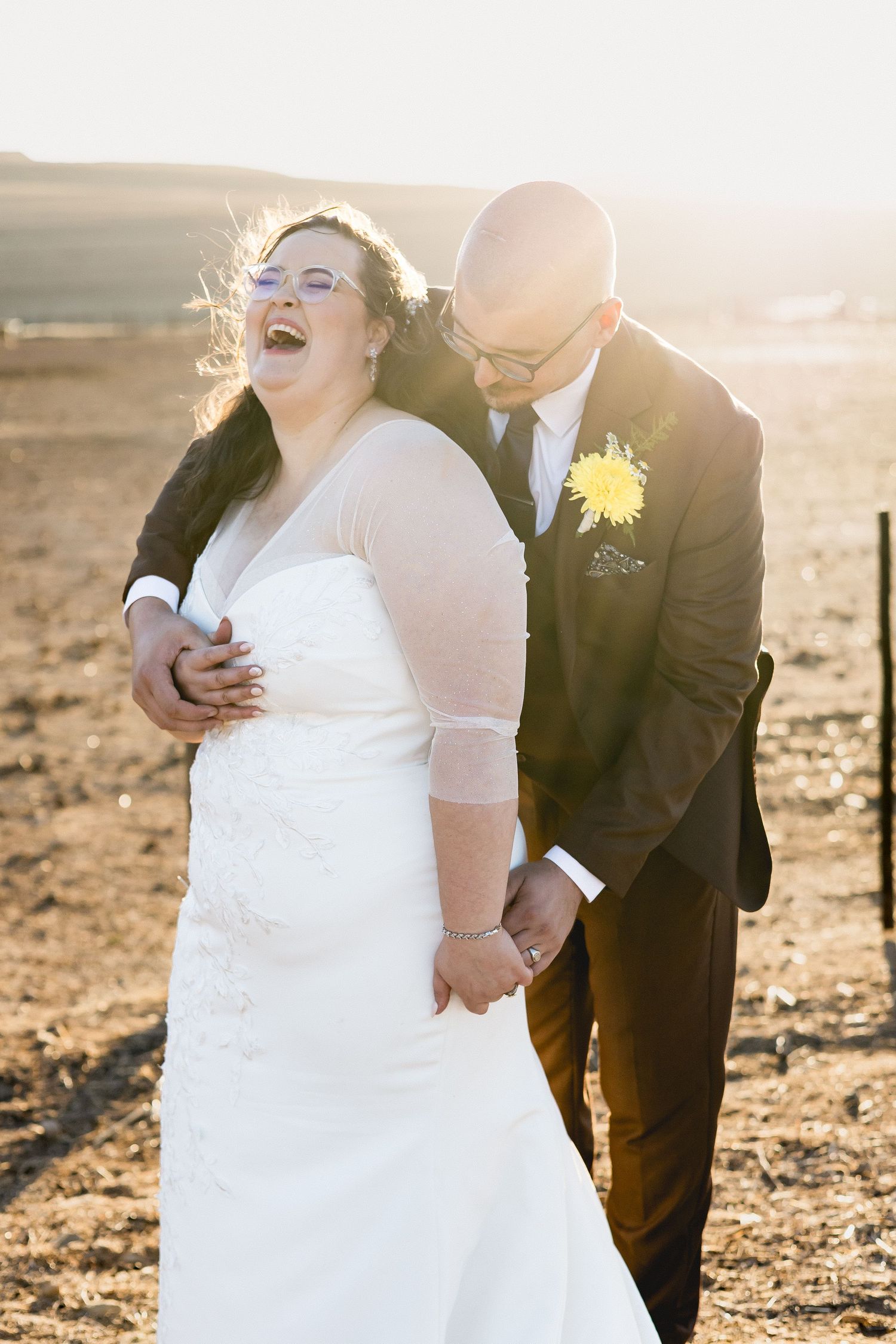 Joyful bride in white gown laughs with bald groom in dark suit and yellow boutonniere at golden hour outdoor wedding.