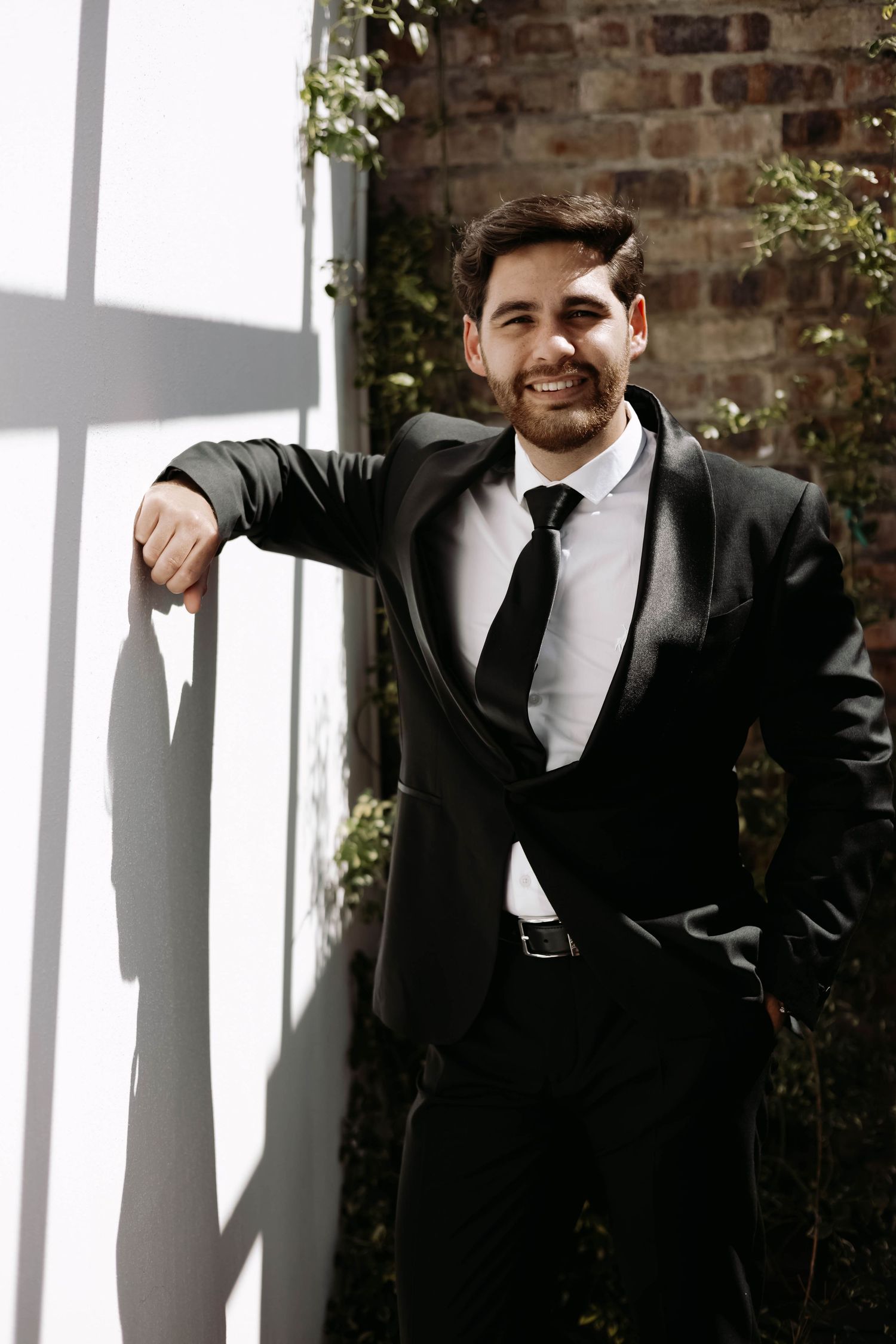 Groomsmen in black suits pose against a rustic brick wall with natural lighting.