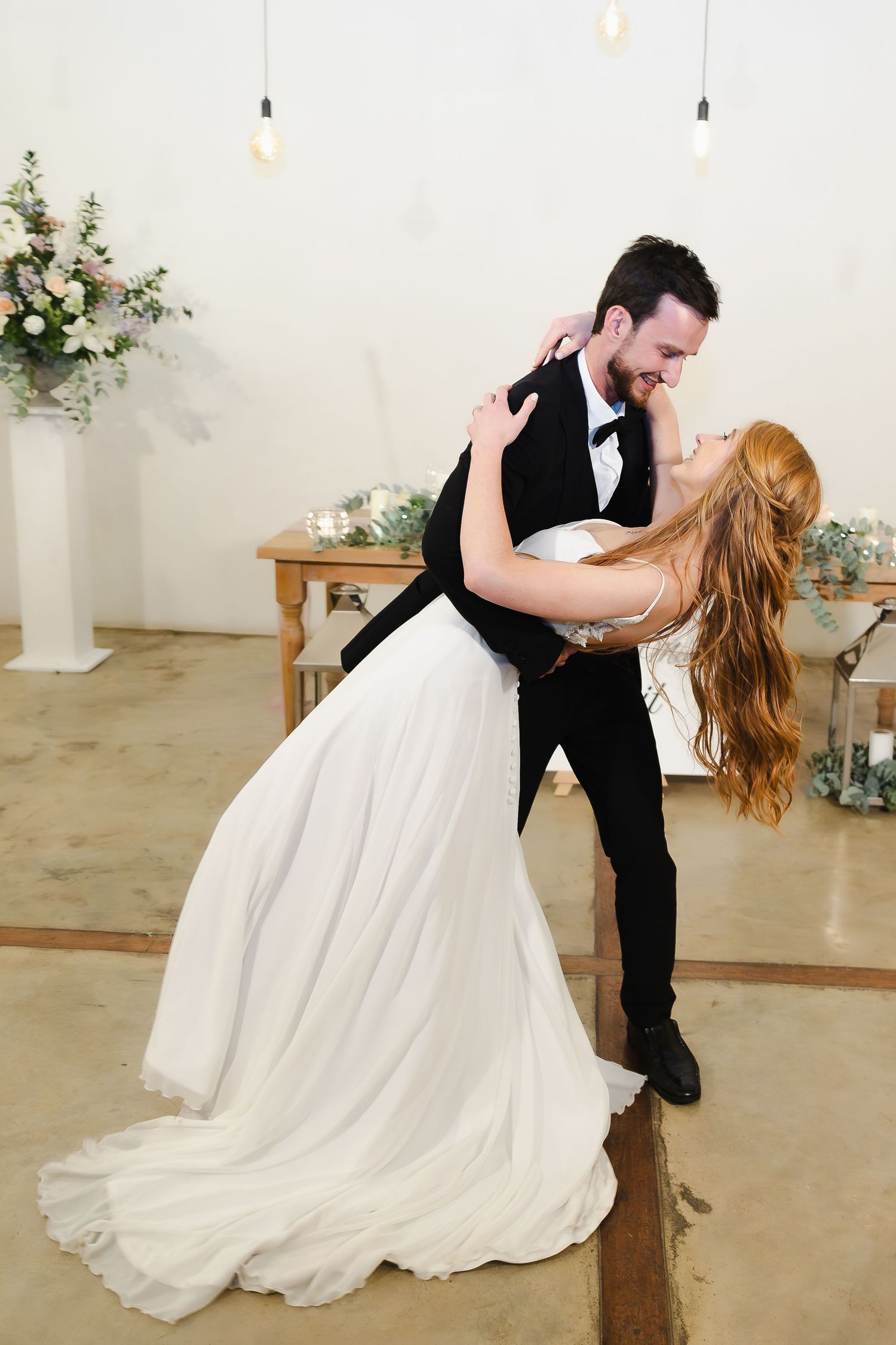 A romantic dip during a first dance between newlyweds in an elegant white venue with floral decorations.