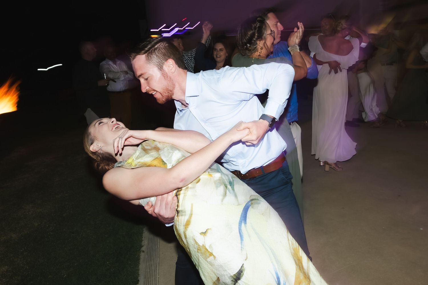 Couples dance together at a dimly lit social gathering with colorful lights in the background.
