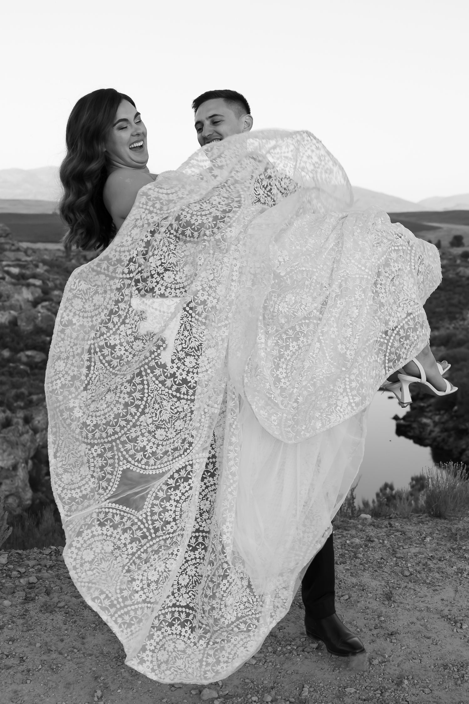 Black and white photo of a joyful couple with the bride's lace dress flowing in the wind.