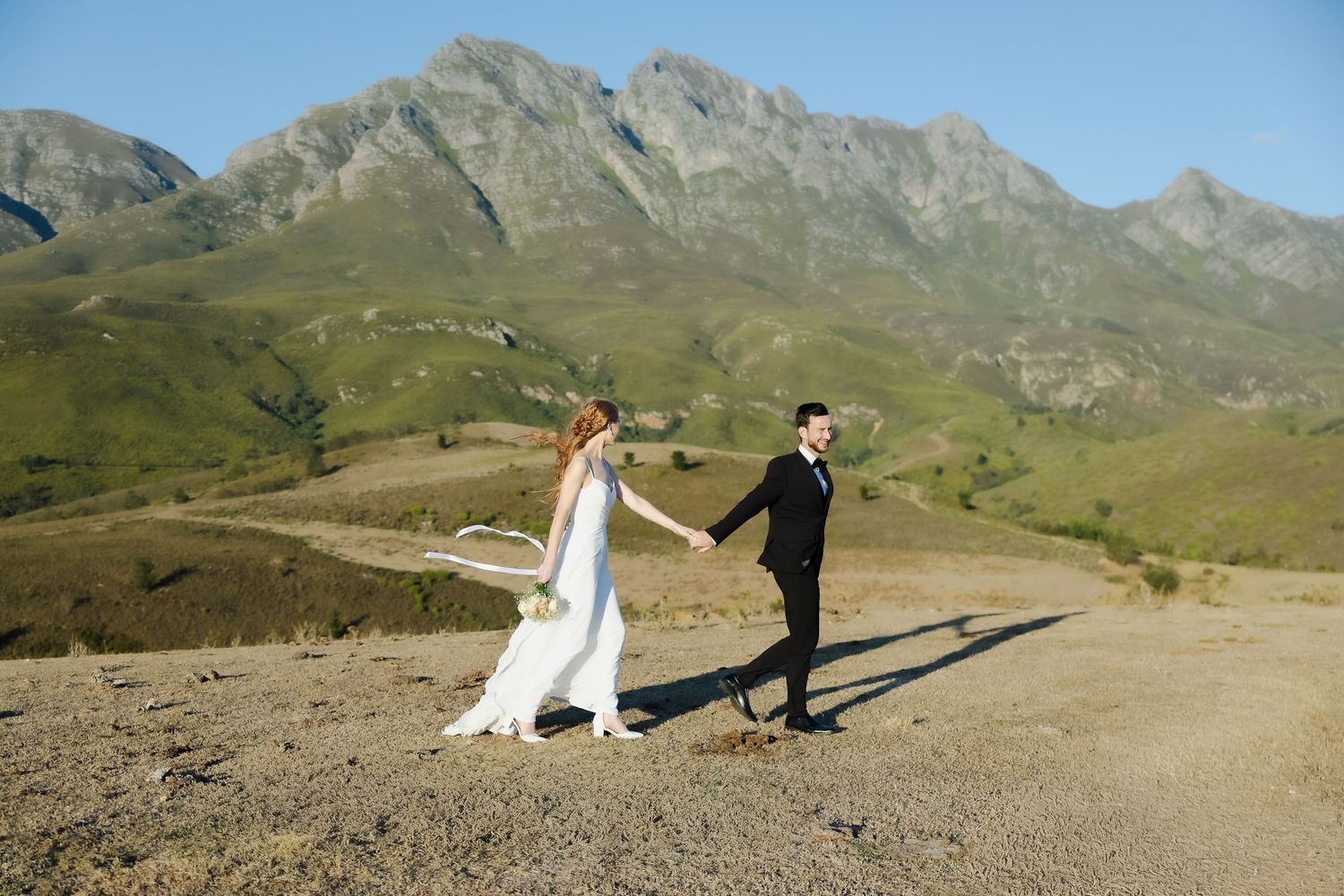 A couple walks along a scenic mountain trail with rugged peaks and green slopes in the background.