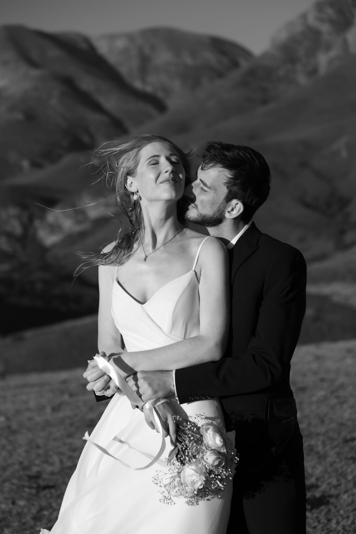 A romantic black and white photo series of a couple in wedding attire embracing against a mountainous backdrop.