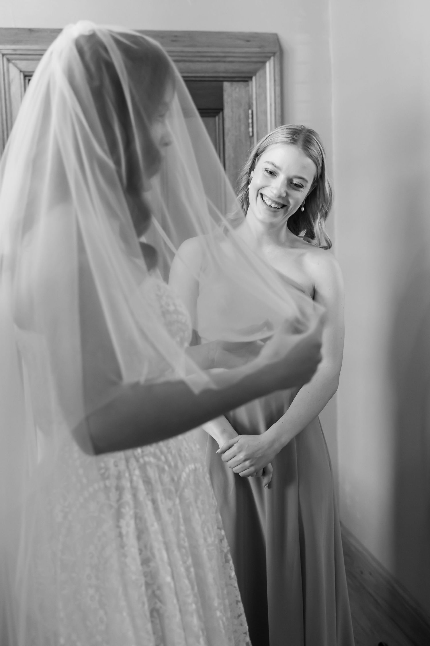 A bridesmaid in a light colored dress smiles while helping with the bride's wedding gown and veil.