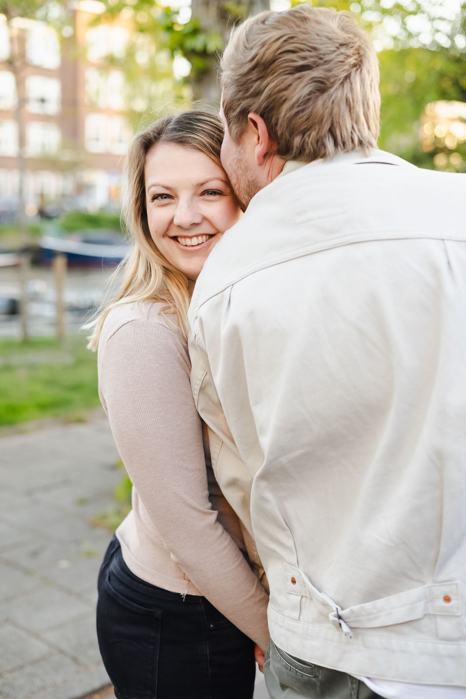 A smiling blonde woman embraces a man outdoors near a tree, looking back at the camera with joy.