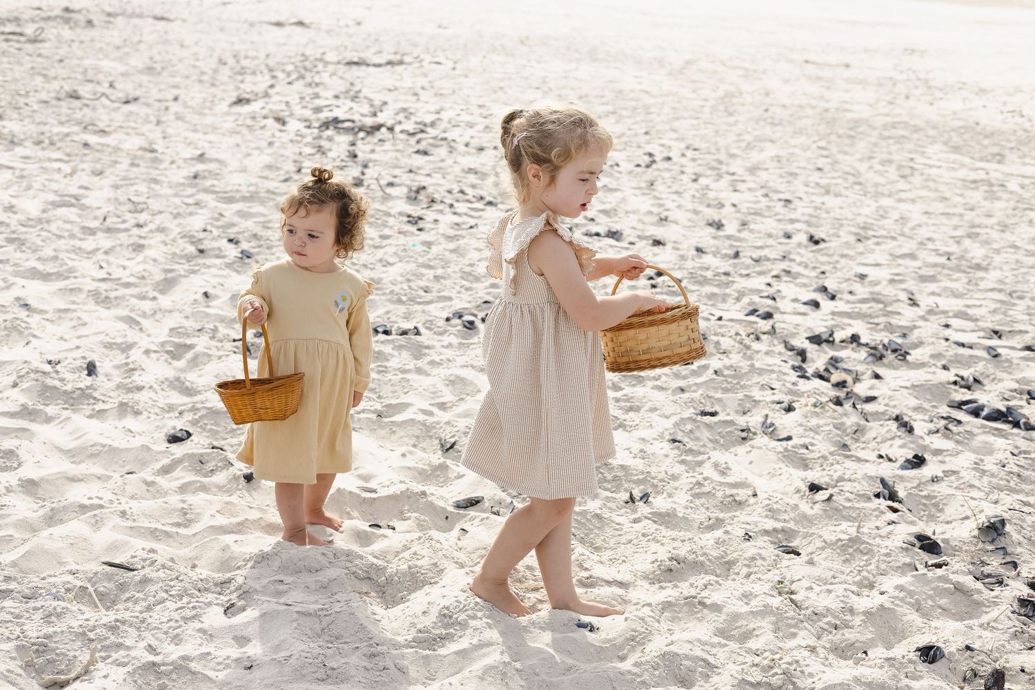 Two young girls in summer dresses carrying wicker baskets while walking barefoot on a sandy beach.