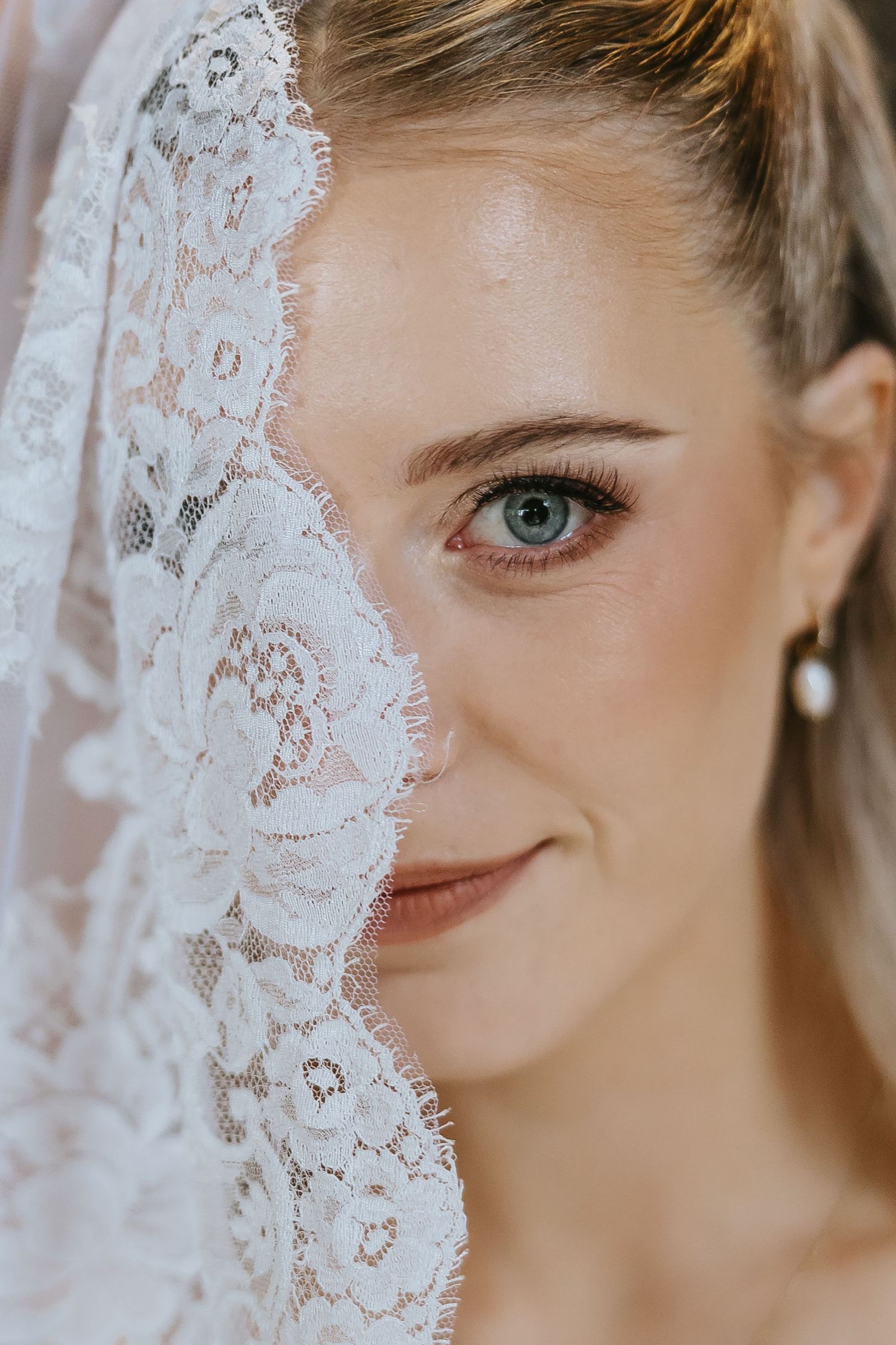 Bride with blue eyes and pearl earring, half face covered by elegant white lace wedding veil, soft bridal makeup.